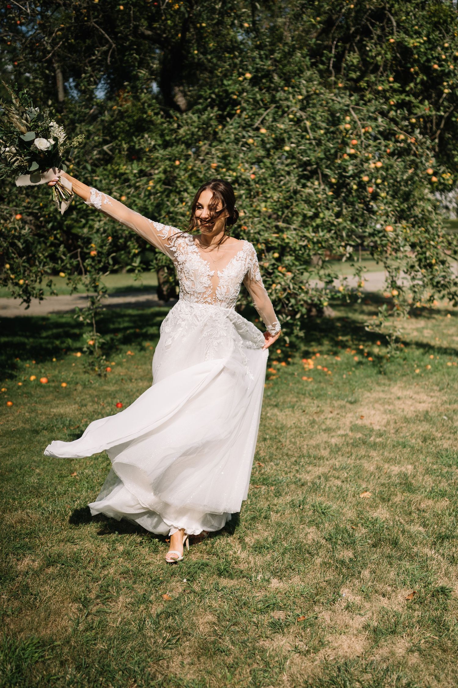 A bride in a lace sleeve wedding gown twirls joyfully in a garden setting.