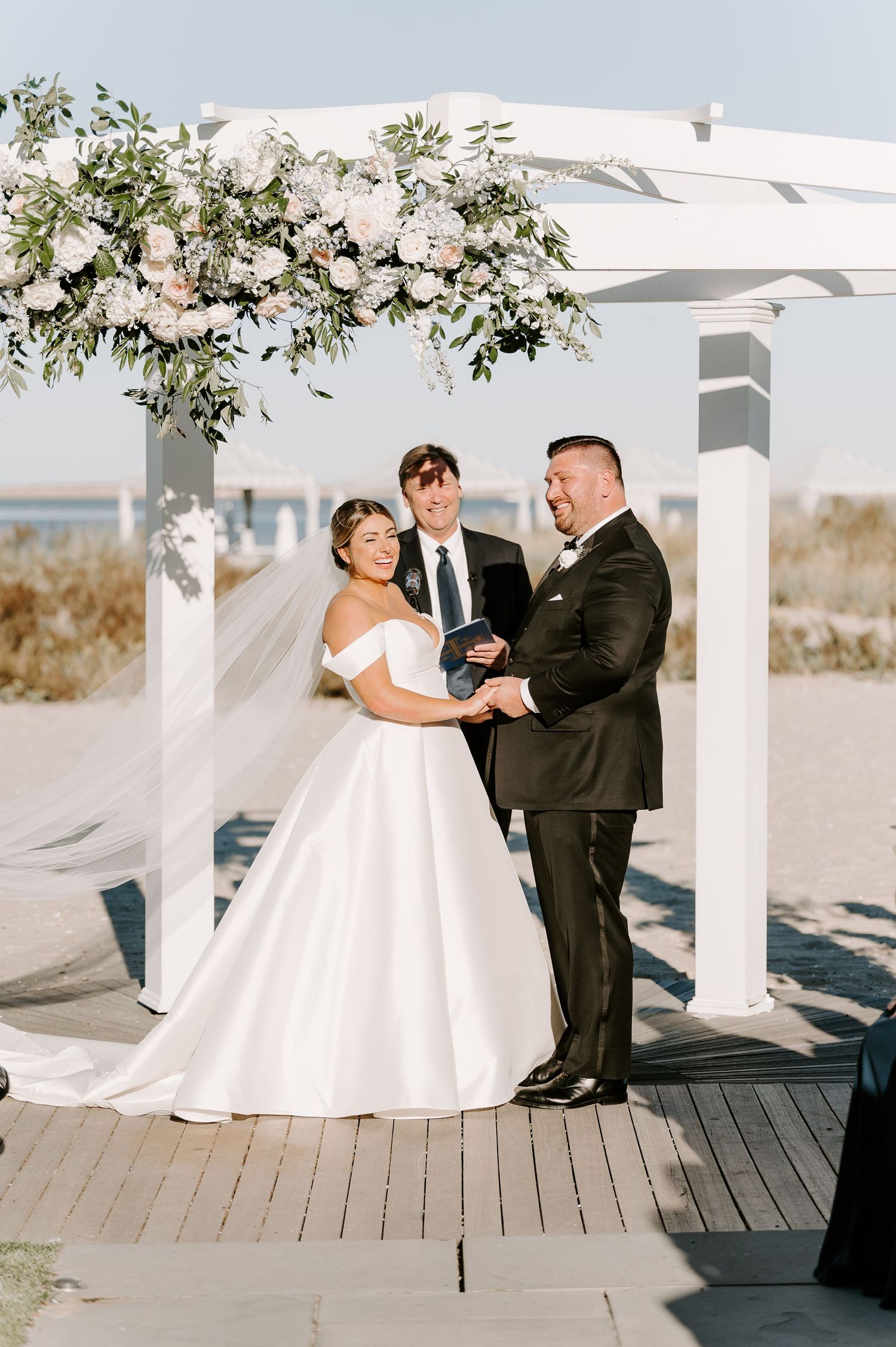 An intimate beach wedding ceremony taking place under a white floral-decorated pergola.