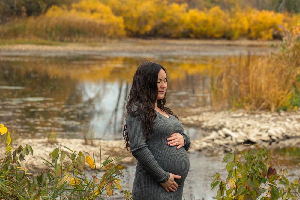 Maternity photo by waterside with golden autumn foliage reflecting in water.