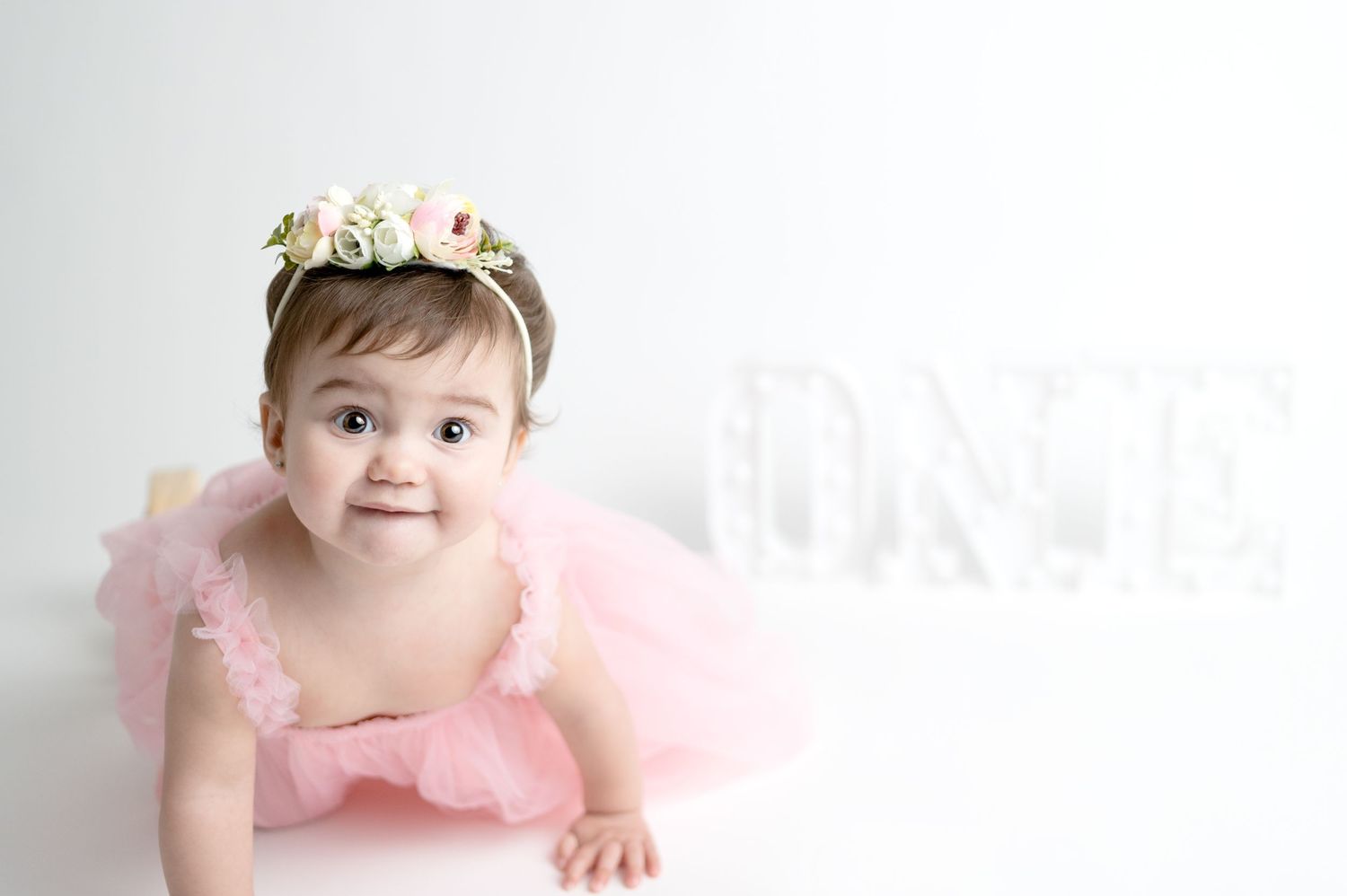 Baby in pink tutu and flower crown crawling on white floor.
