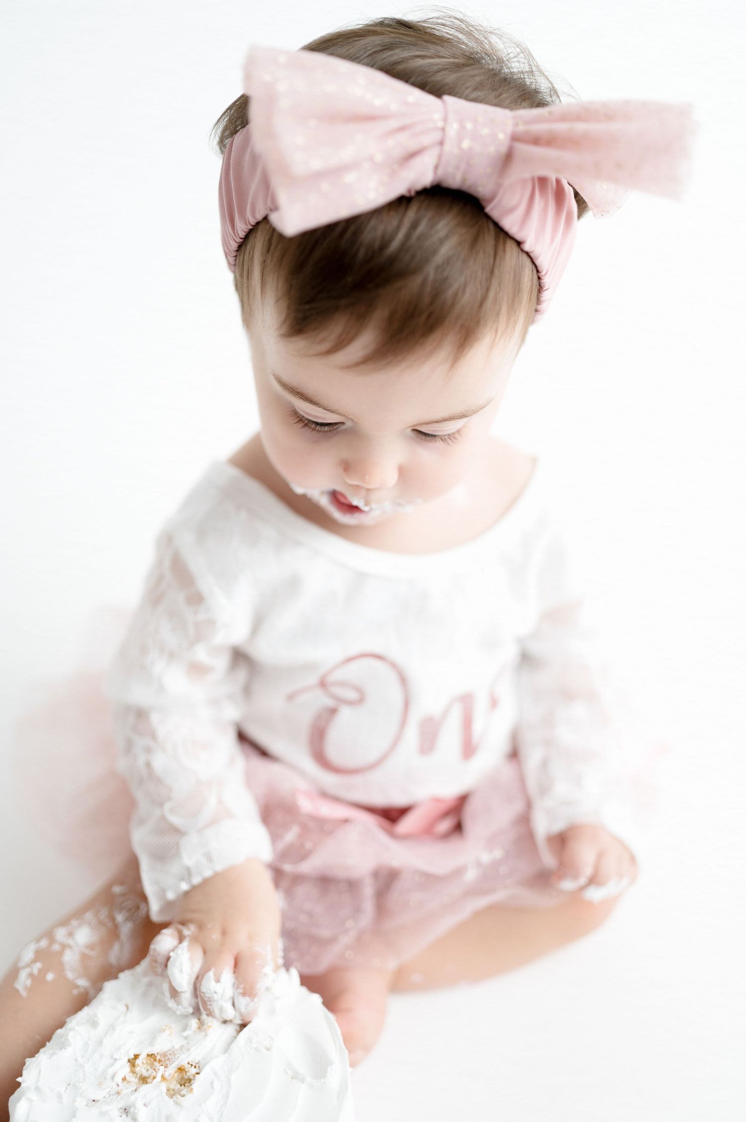 Baby in pink bow headband and white dress looking down at floor.