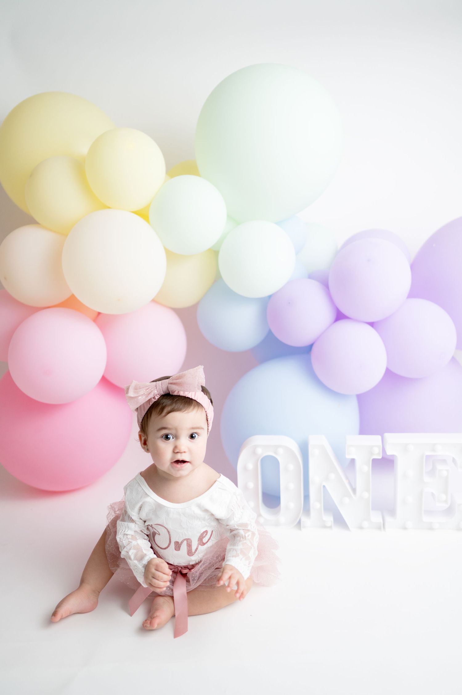 First birthday photo setup with pastel balloon arch and white ONE letters on white background.