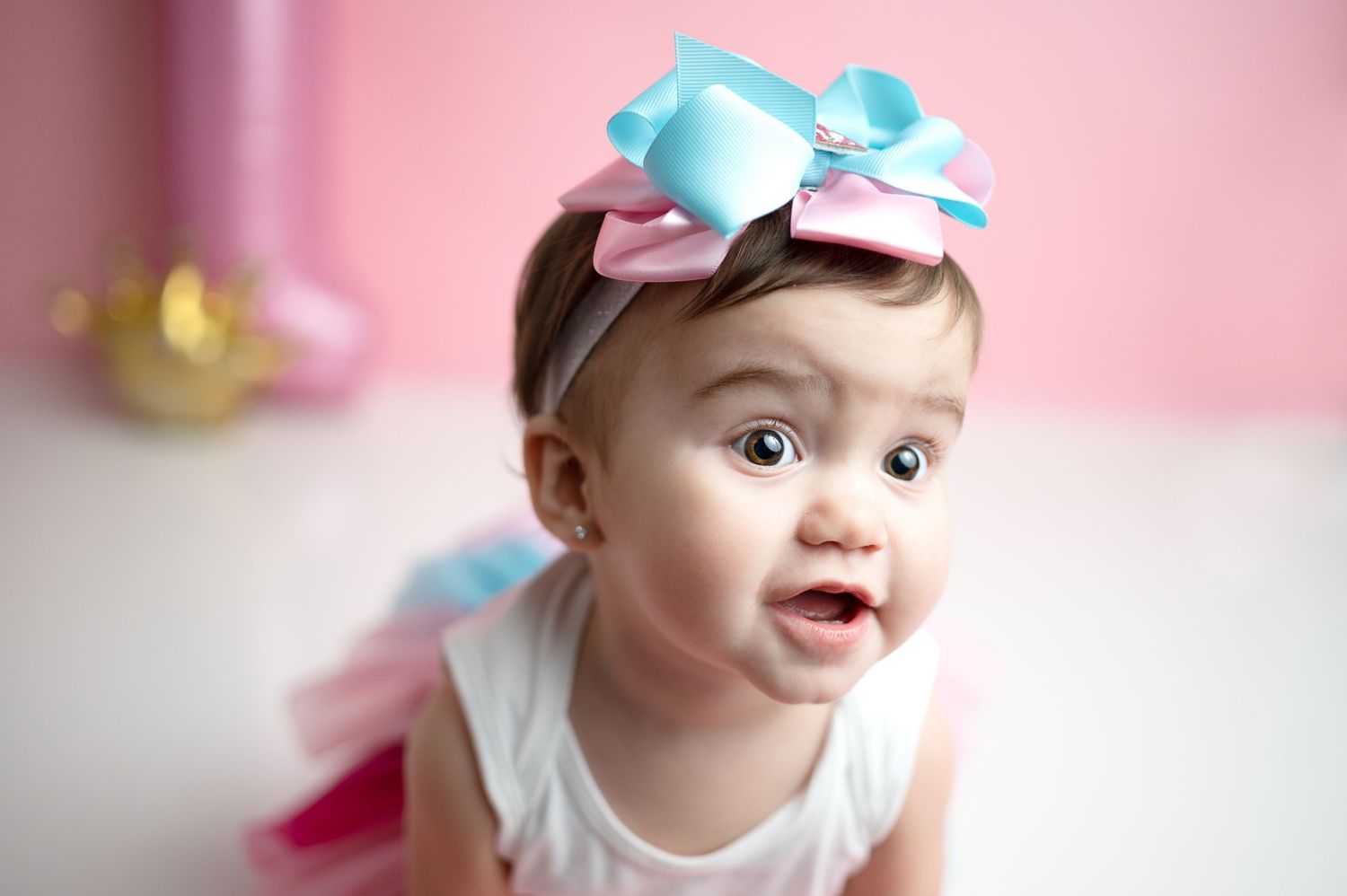 Baby wearing a blue and pink bow headband against a pink background with joyful expression.