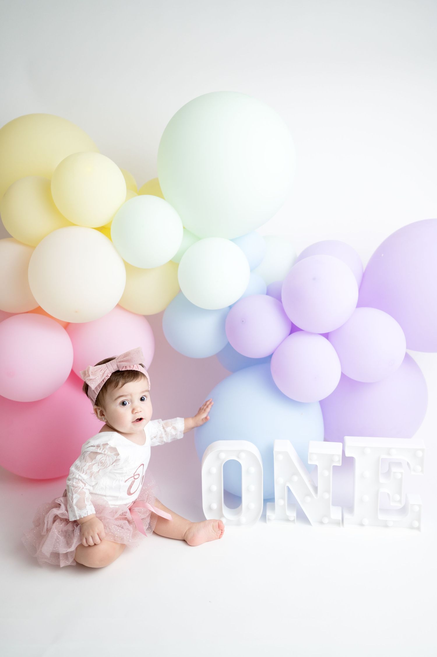 Baby in tutu reaching toward white ONE letters with pastel balloon backdrop.
