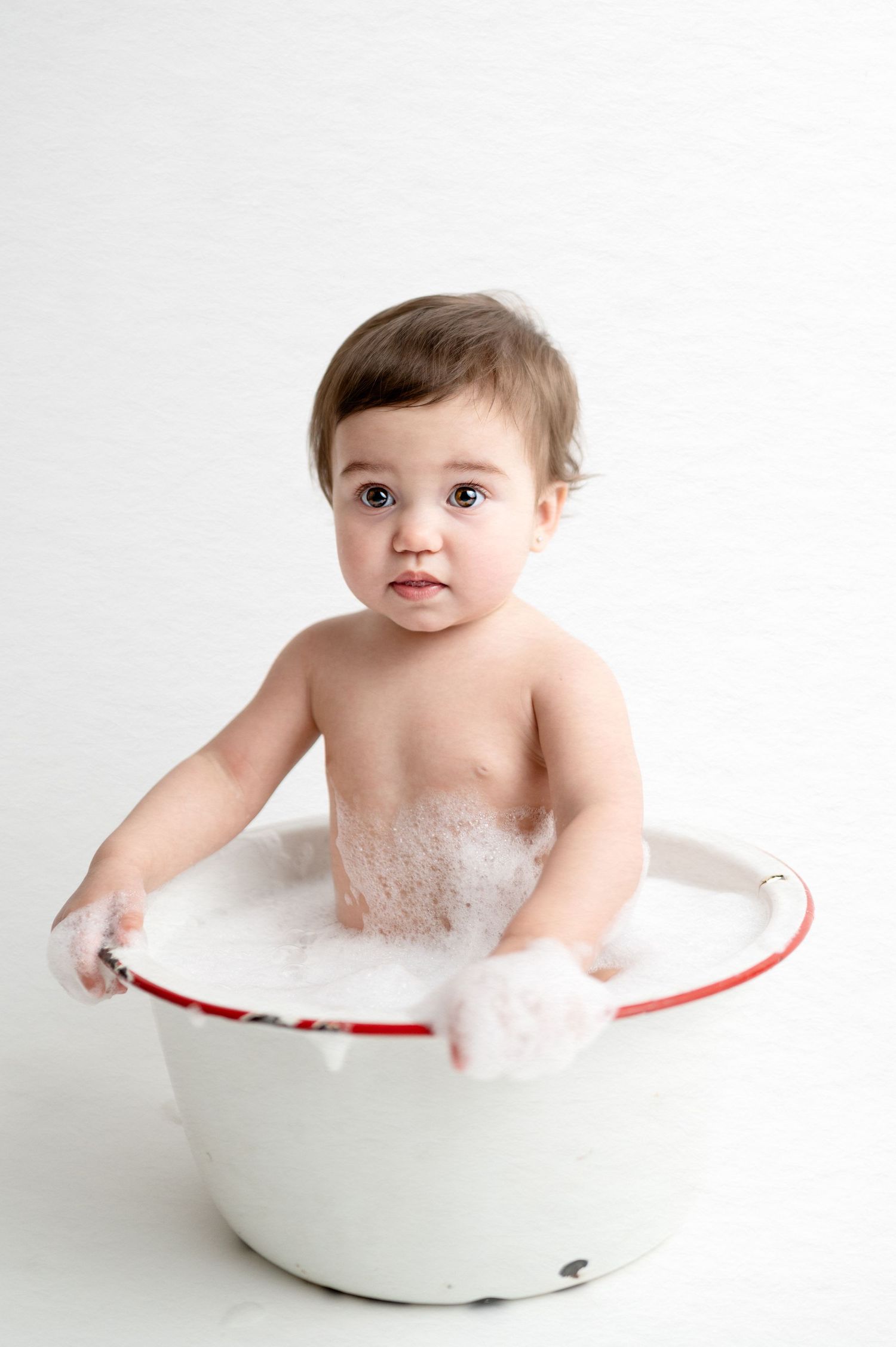 Baby enjoying bath time in white enamel tub against white background.