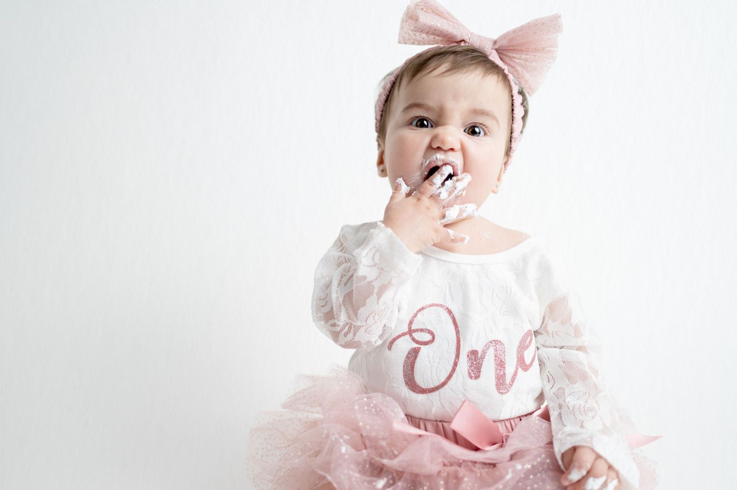Baby wearing pink bow tasting birthday cake frosting with messy hands.