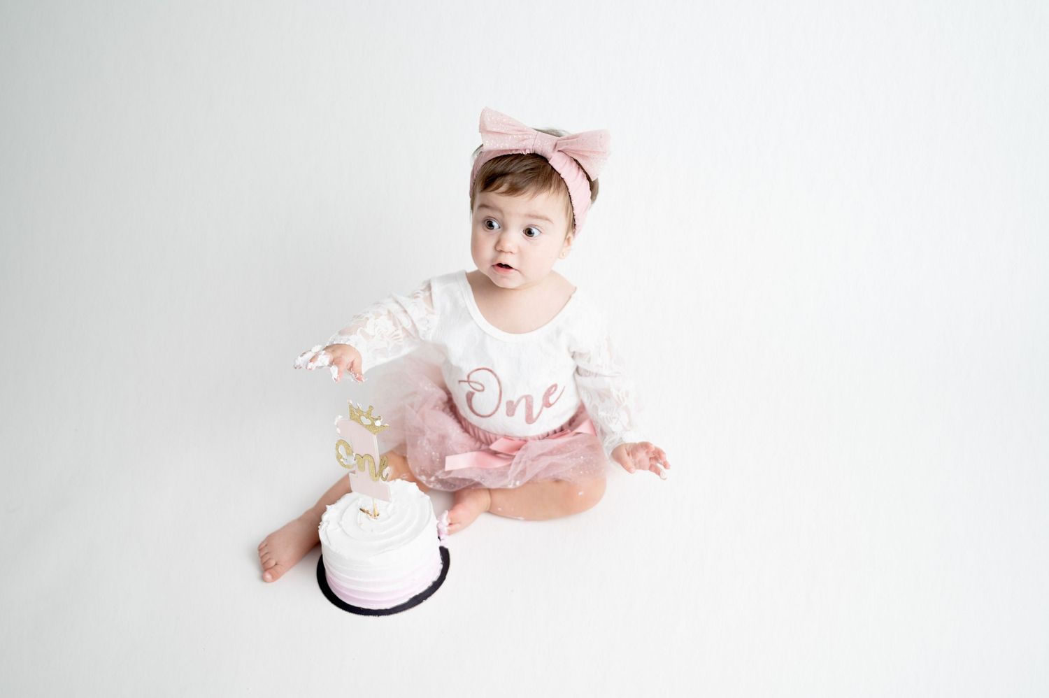 Baby in white dress and pink bow sitting with small cake on white background.