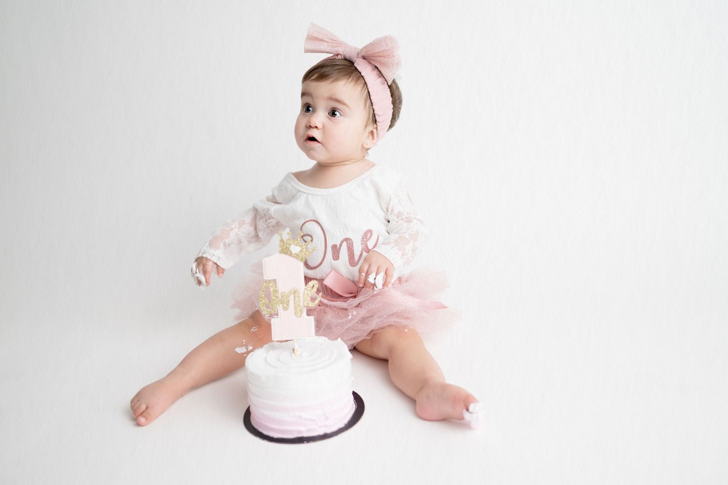 Baby in white outfit with pink bow sitting next to small white birthday cake.