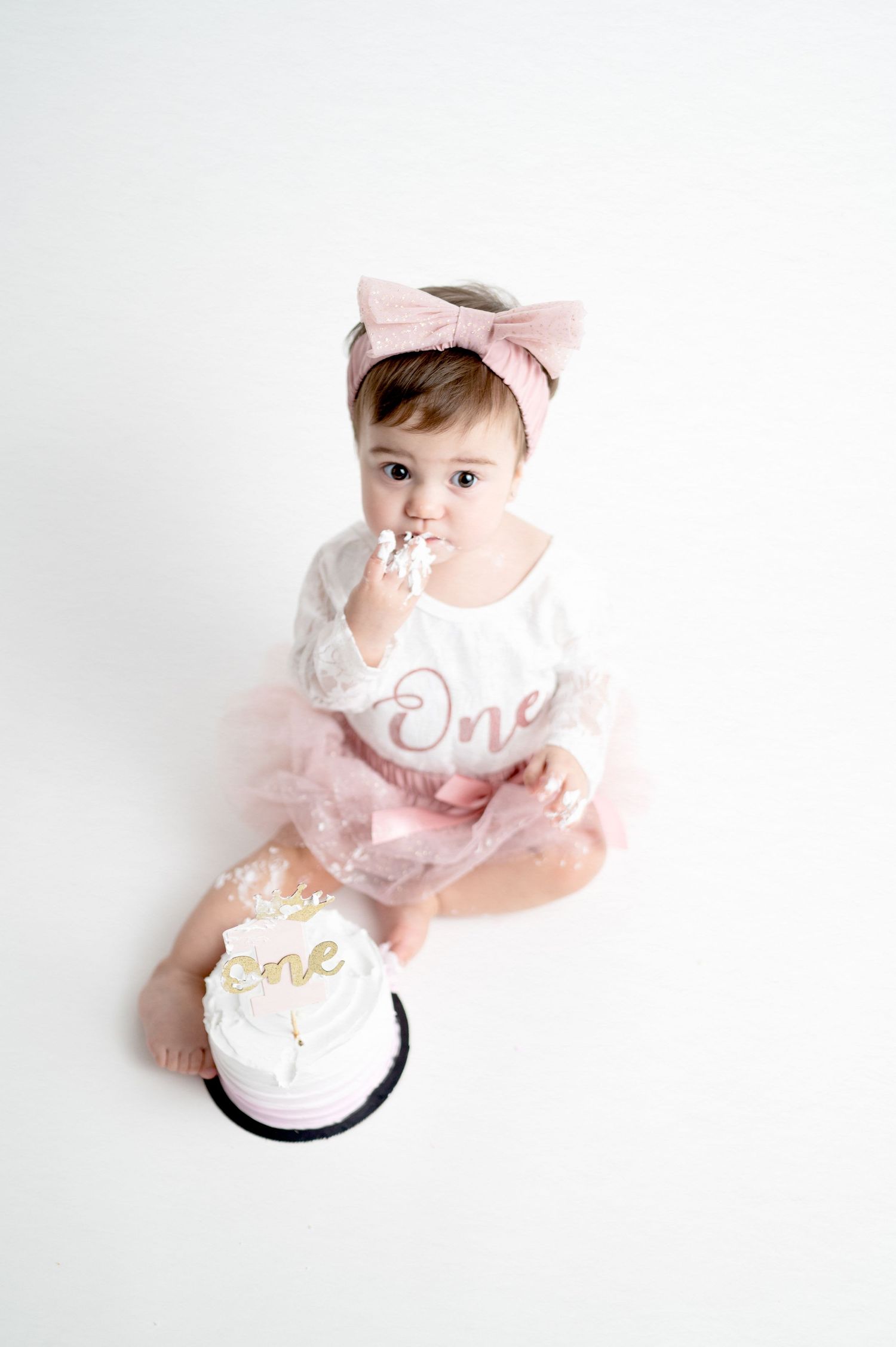 Baby in tulle dress eating cake while sitting on white background.