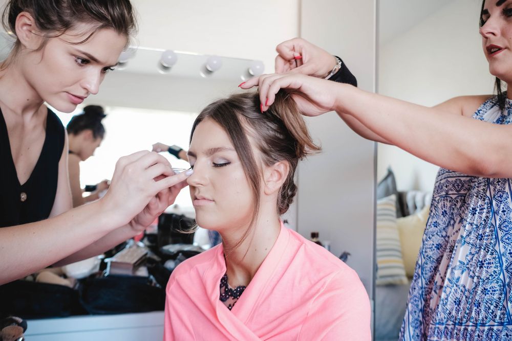 Makeup artists work on styling in a bright salon setting with vanity lights and mirrors.