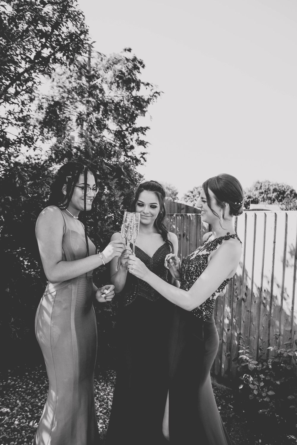 Black and white photo of three friends in elegant dresses toasting at an outdoor gathering.