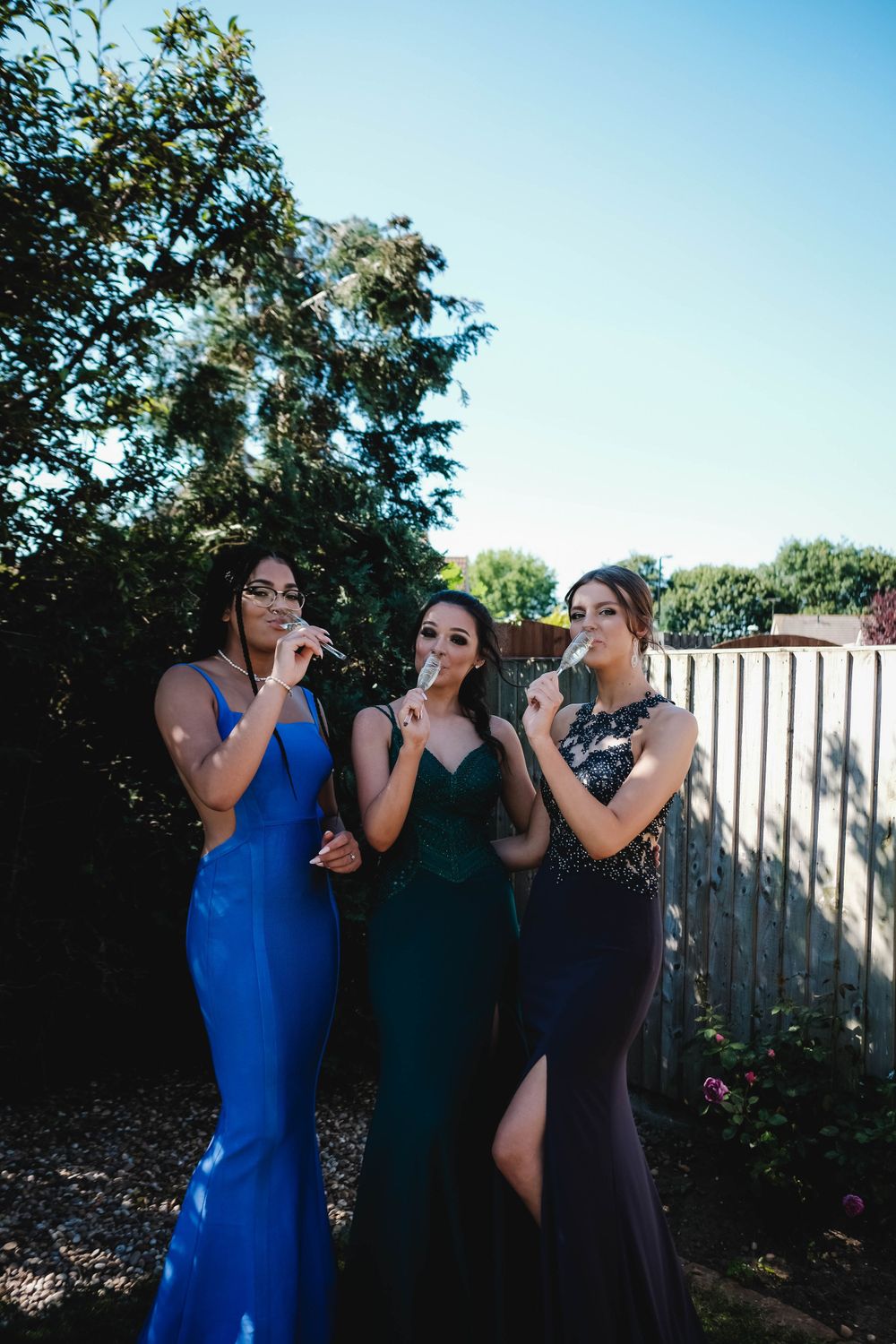Three friends in elegant formal dresses pose playfully together in a backyard garden setting.