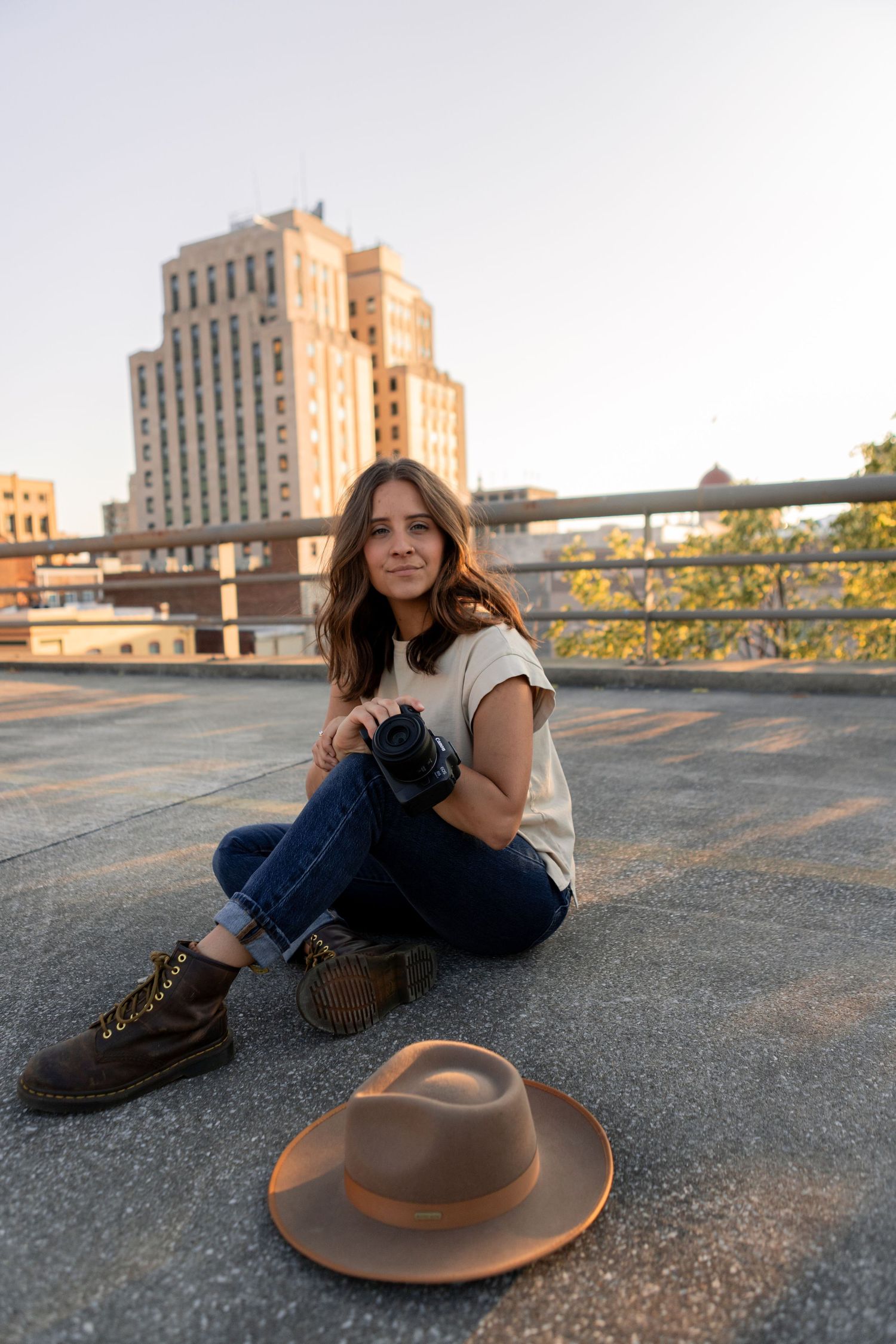 Person in jeans and white shirt lounging on rooftop with brown hat and city buildings in background.