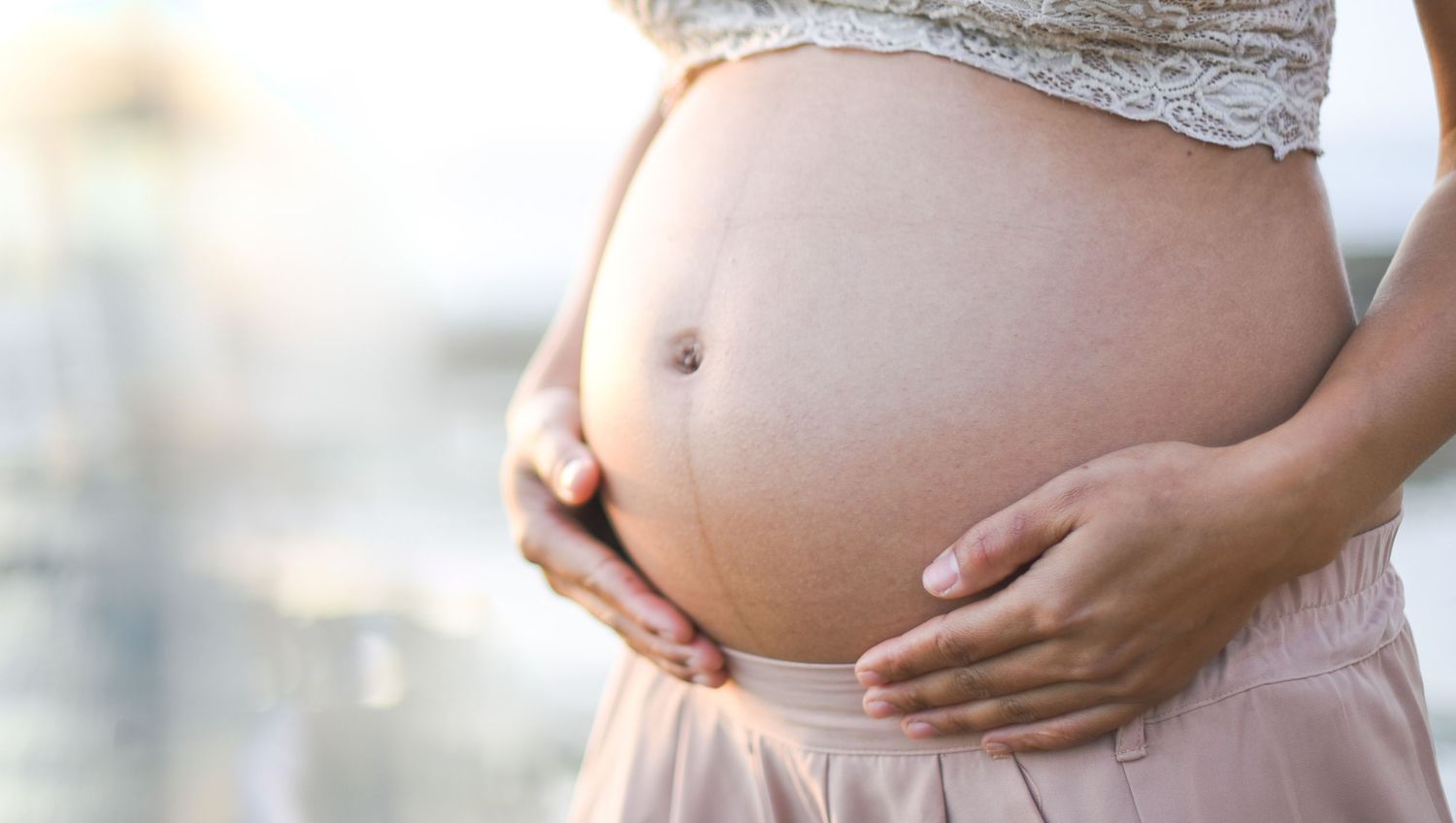 Close up of pregnant belly with hands cradling bump against blurred background.
