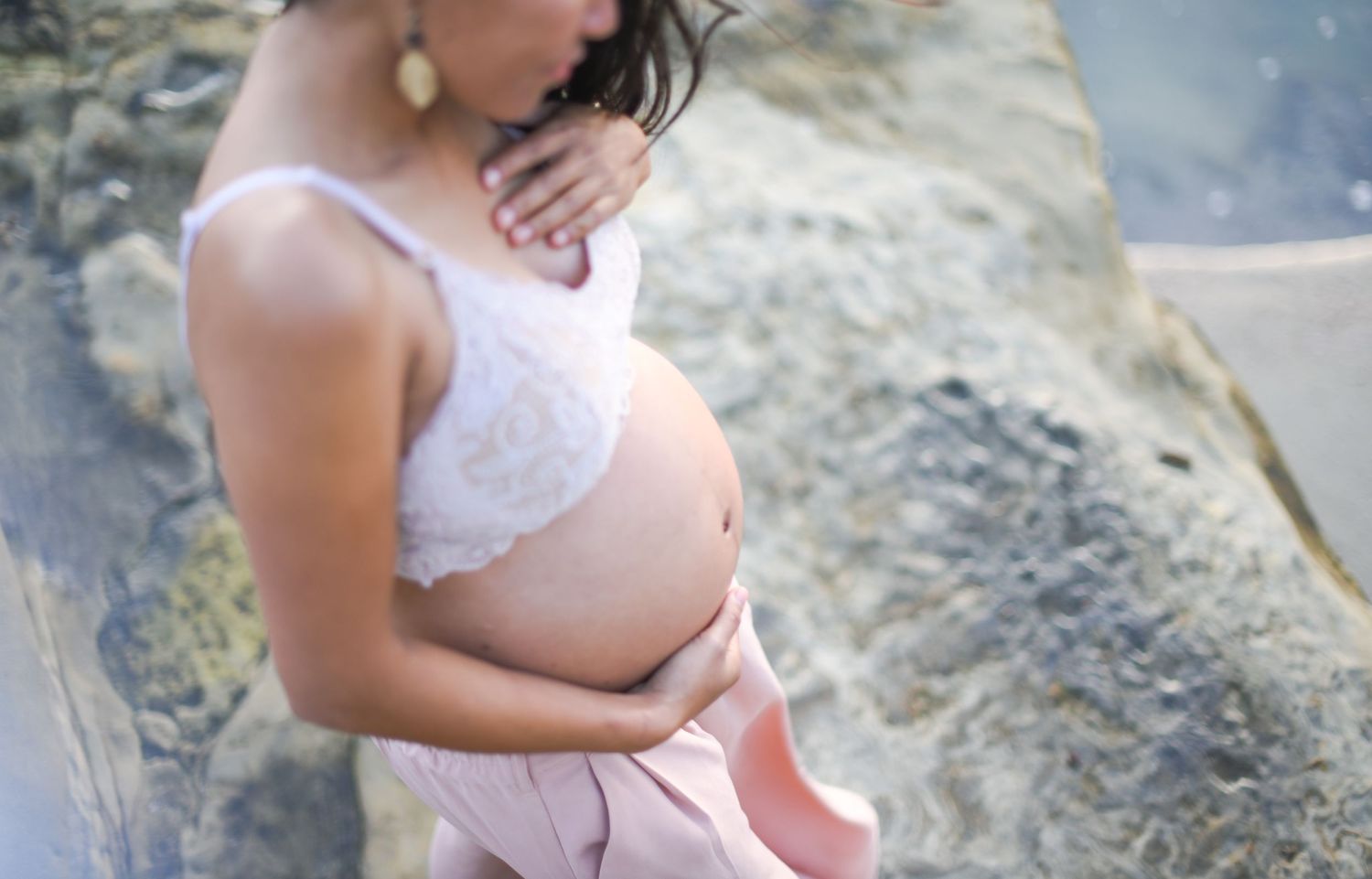 Maternity silhouette in white lace dress against rocky coastal backdrop.