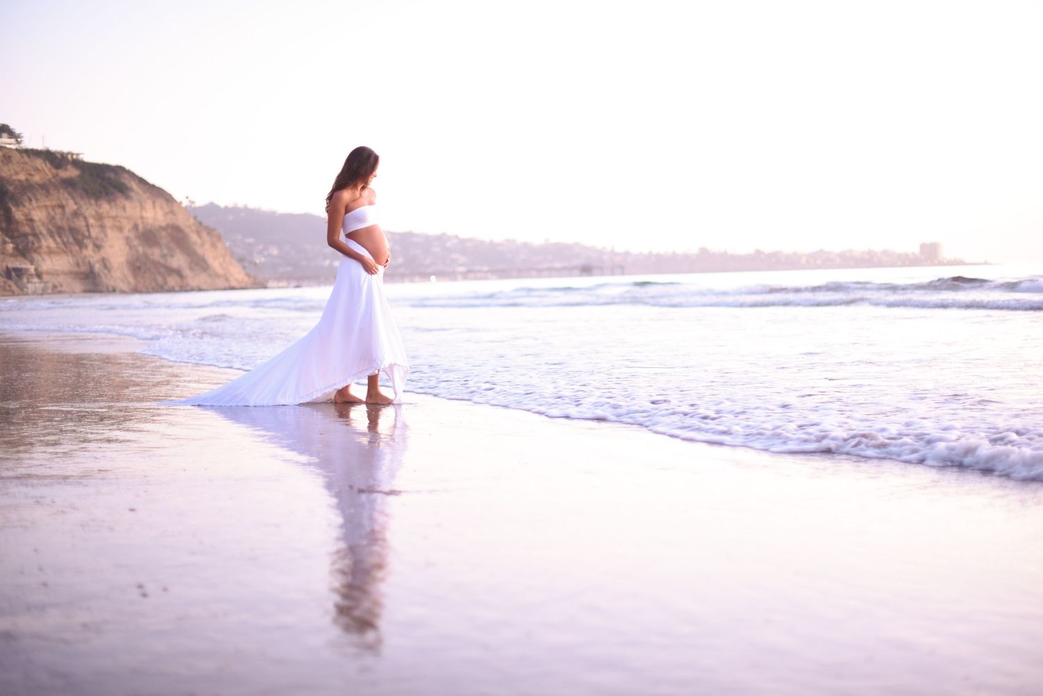 Woman in flowing white dress stands at waters edge during sunset on beach.