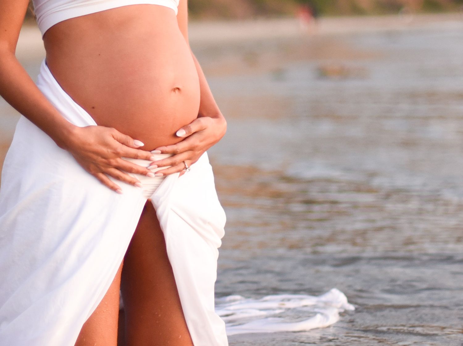 Expectant mother in white dress cradling baby bump on beach at sunset.