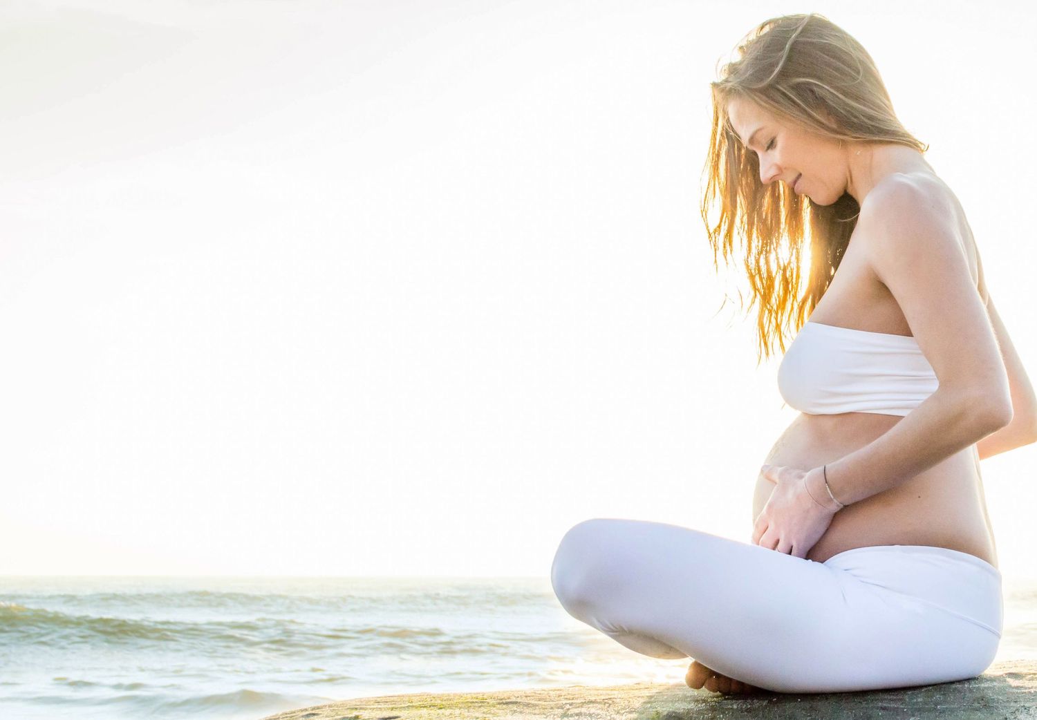 Pregnant woman in white clothing meditating by ocean at sunset.