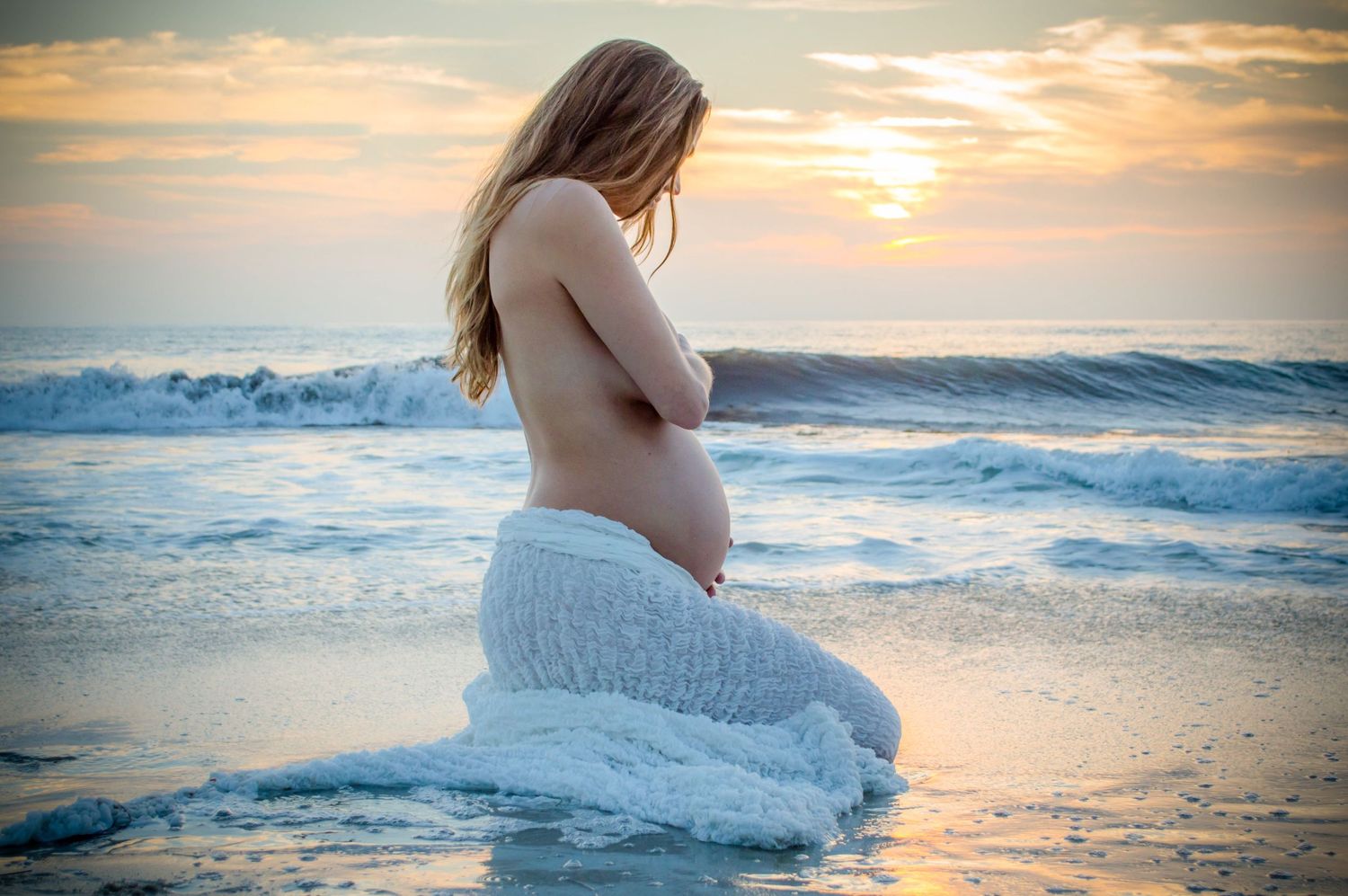 Silhouette of pregnant woman kneeling in white dress at beach during golden sunset.
