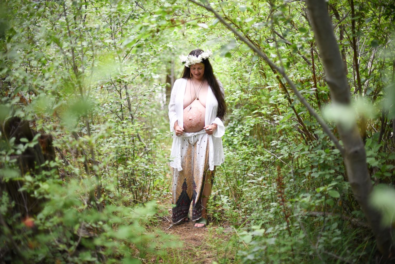 A maternity photo shoot in a lush green forest path shows an expecting mother in a flowing white dress.