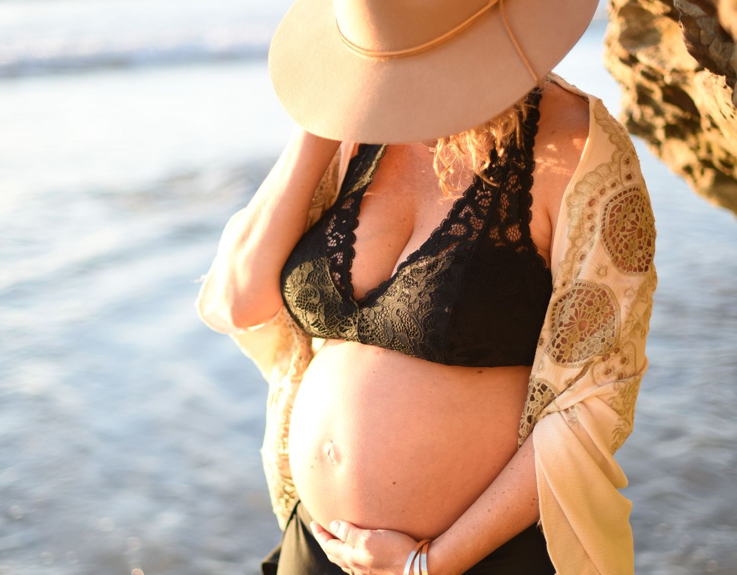 Close-up of pregnant belly with black lace bralette and floppy hat at the beach.