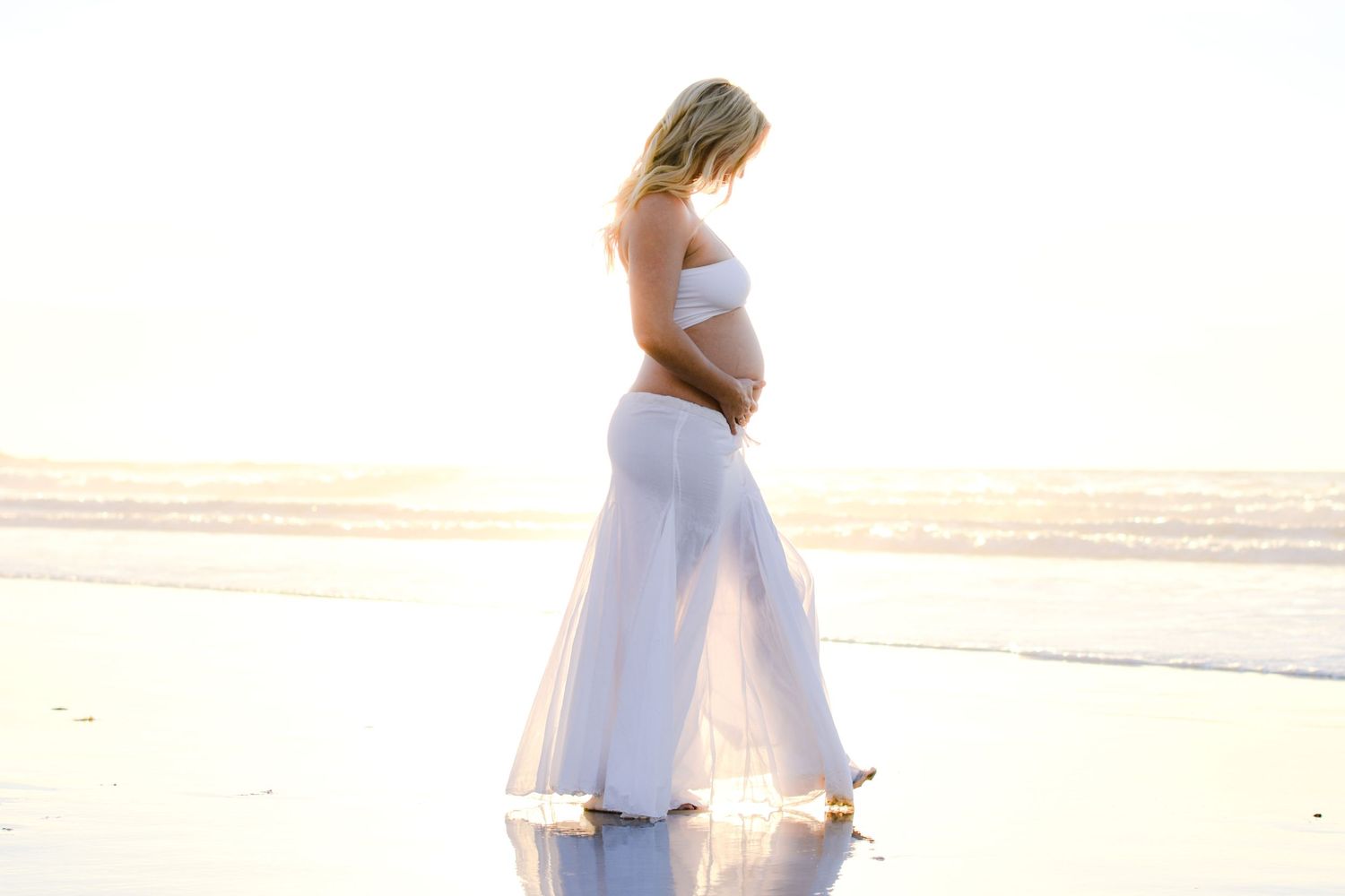 Silhouette of a pregnant woman in white dress standing on reflective beach at sunset.