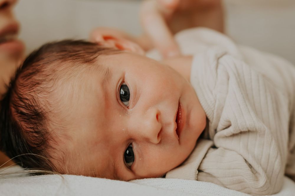 Close up of baby lying on white bedding with big bright eyes looking at camera.