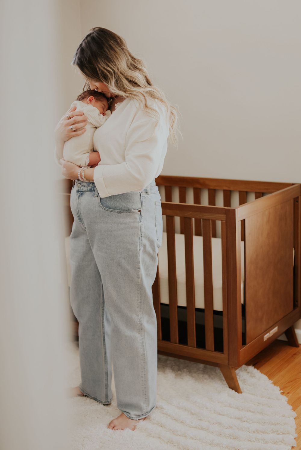Parent in white sweater and jeans holding baby near wooden crib in nursery with round white rug.