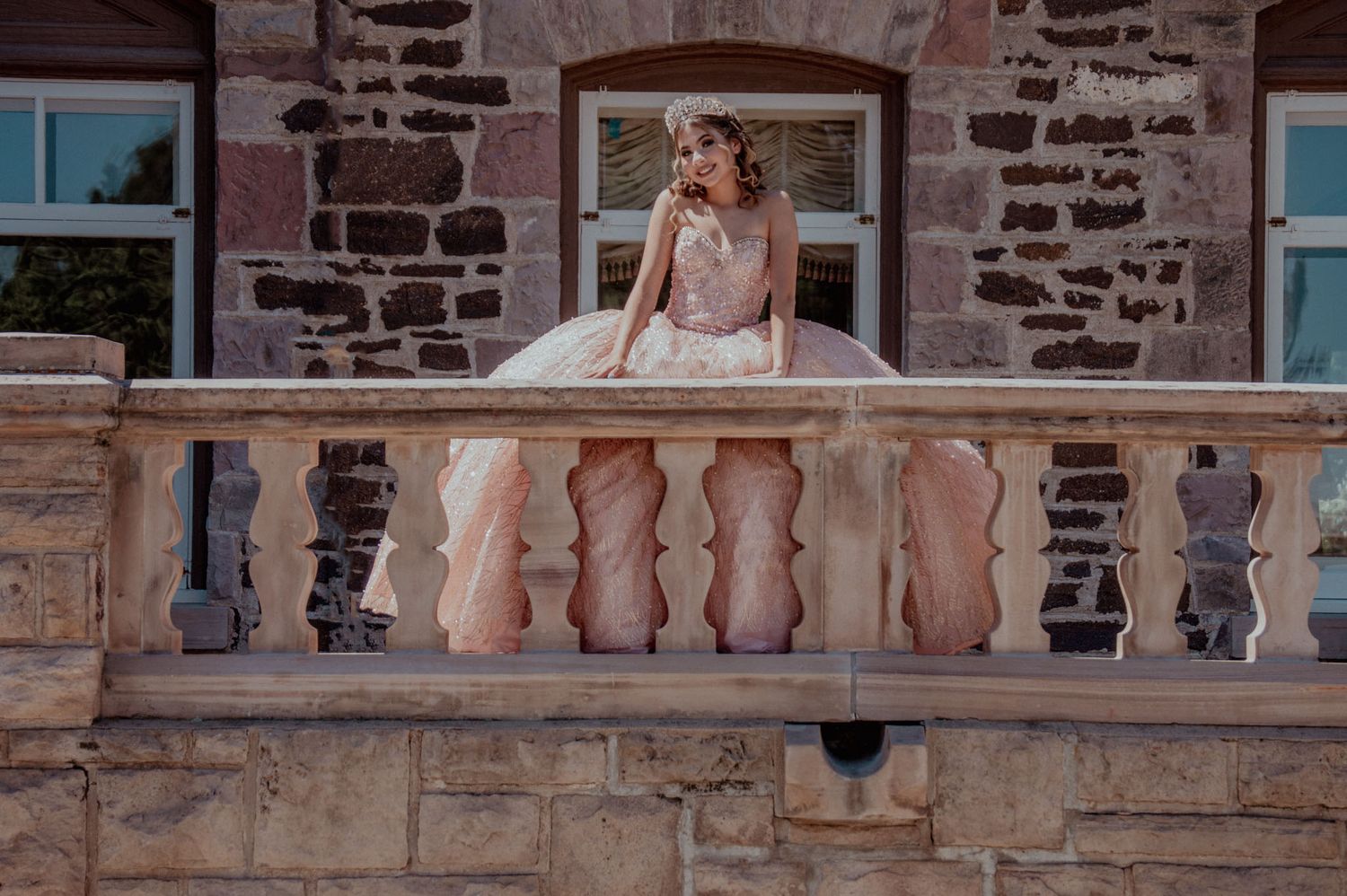 Elegant woman in pink ballgown posing on stone balcony of historic building.