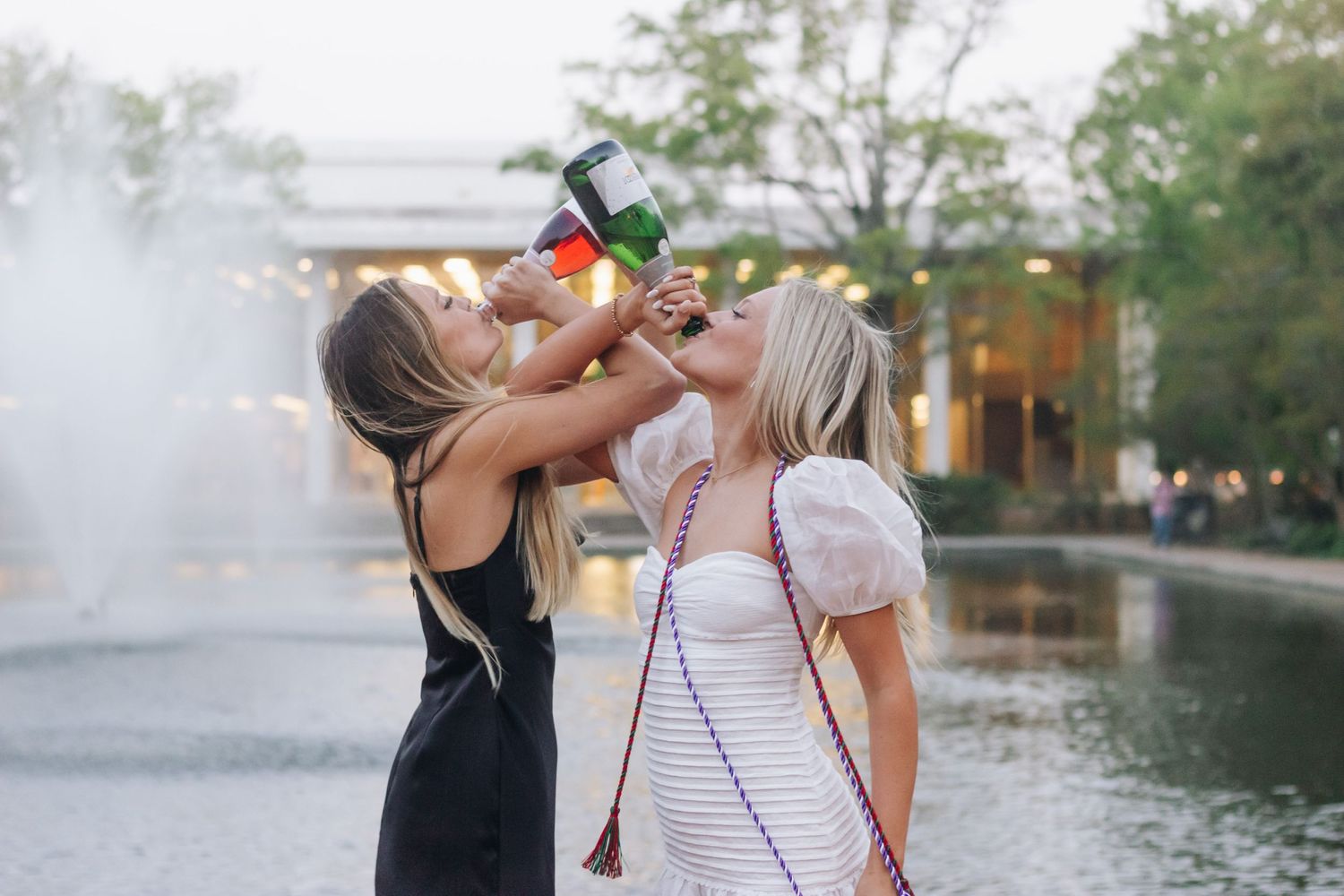 Two graduates celebrate by fountain with one spraying champagne.
