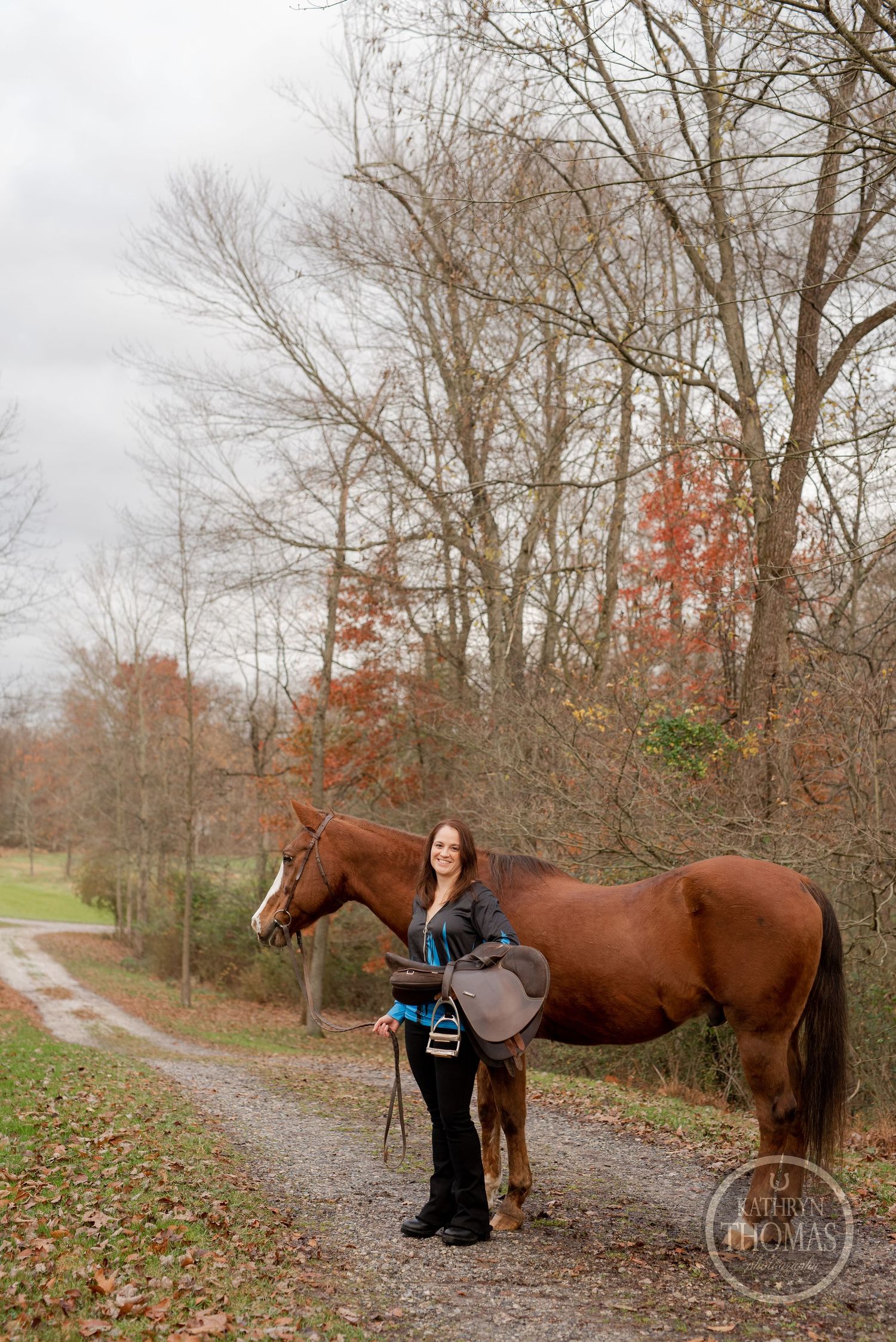 A chestnut horse and companion stand on a country path with fall foliage and a red barn in the background.