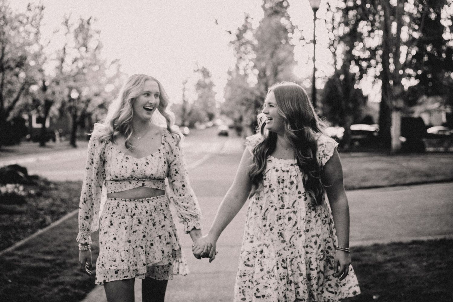 Two friends in floral dresses hold hands while walking down a path in black and white.