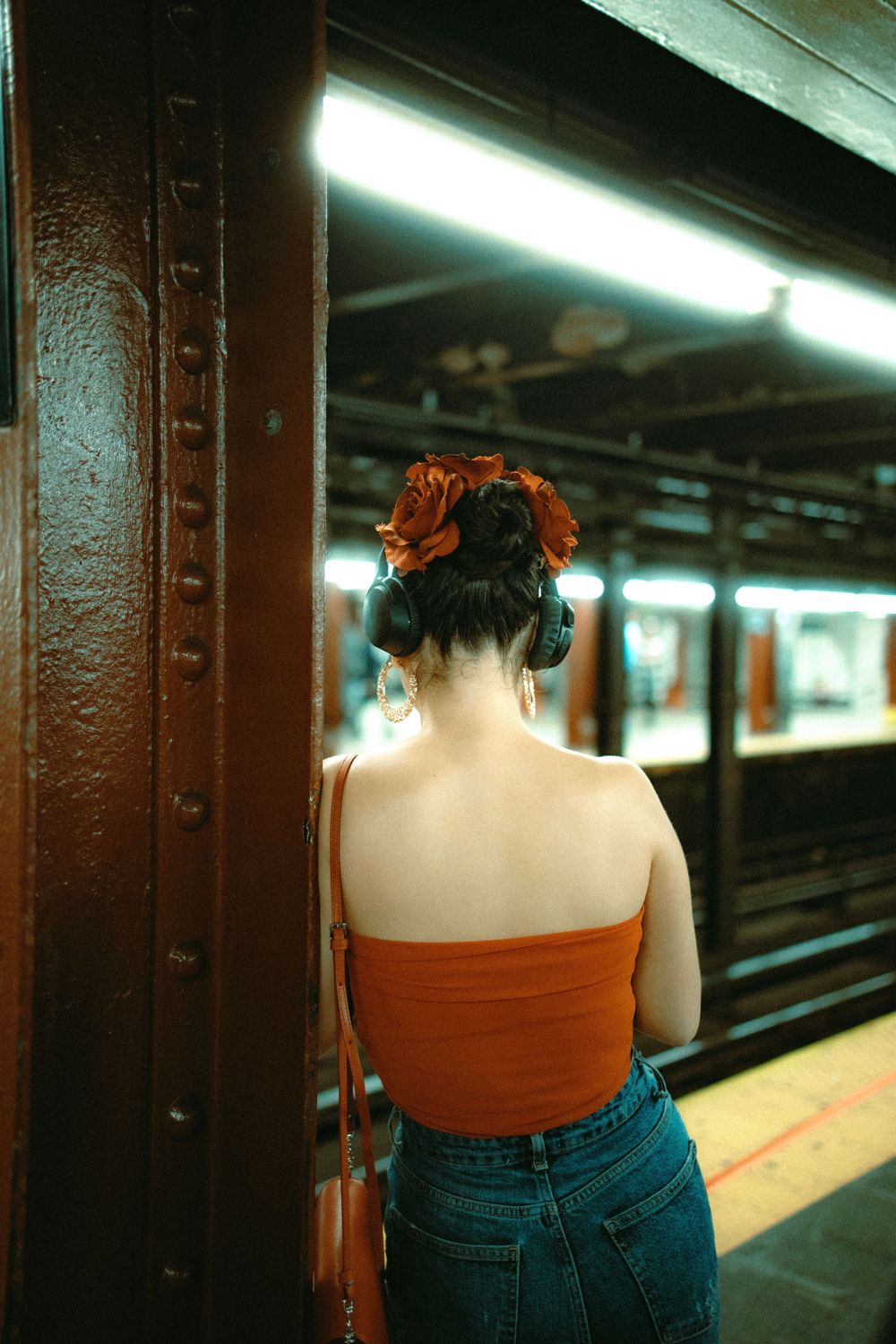 Woman in orange top and jeans standing at a subway platform with red-dyed hair.