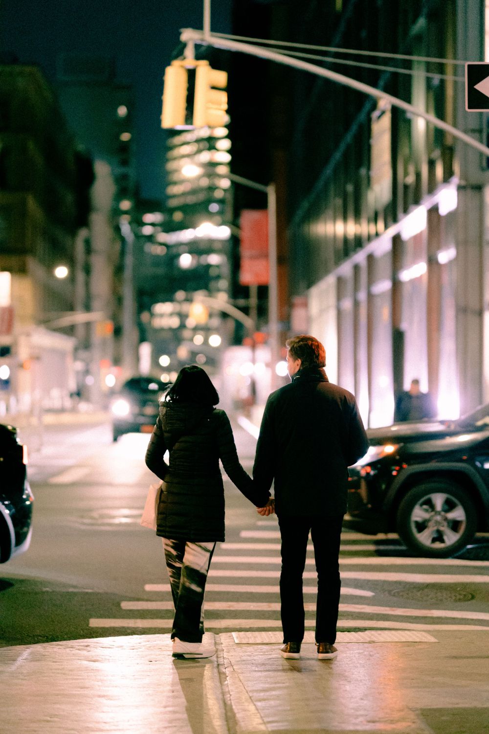 Two figures holding hands walk down a dimly lit city street at night.