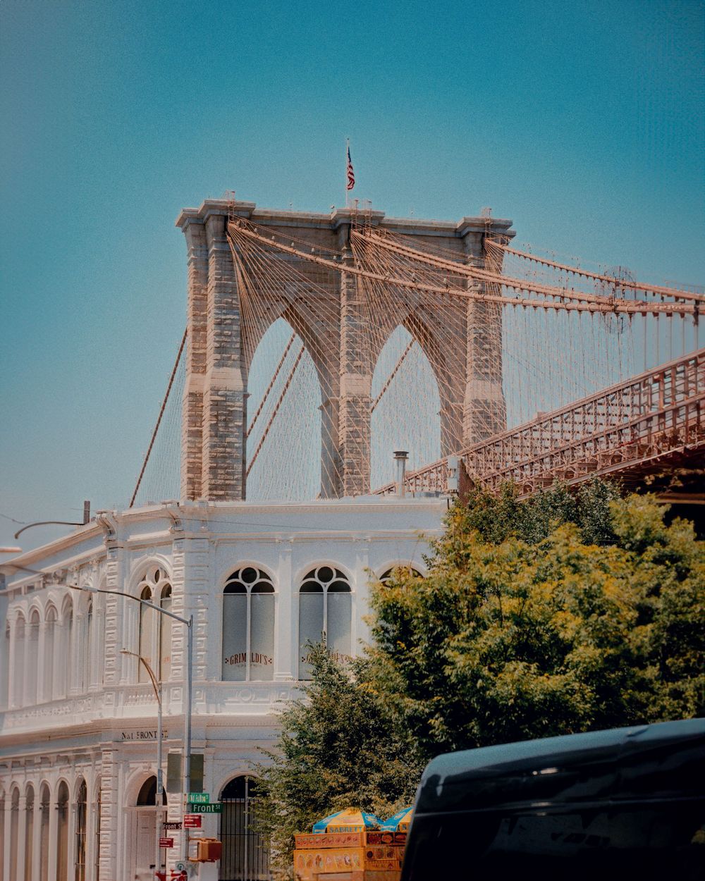 The iconic Brooklyn Bridge stone arch and suspension cables rise against a bright blue sky.