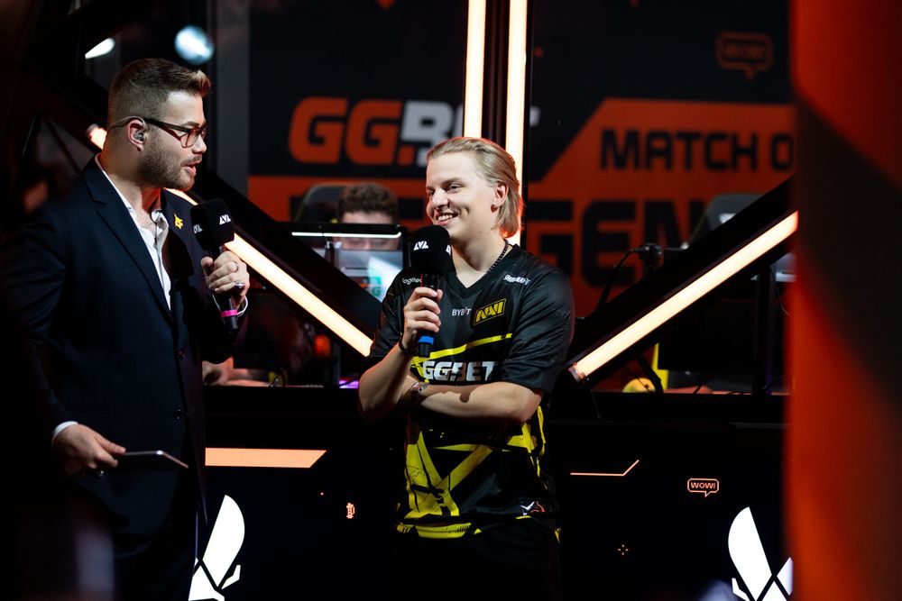 Two esports commentators on stage at a gaming event with yellow and red lighting and GG branding visible in background.