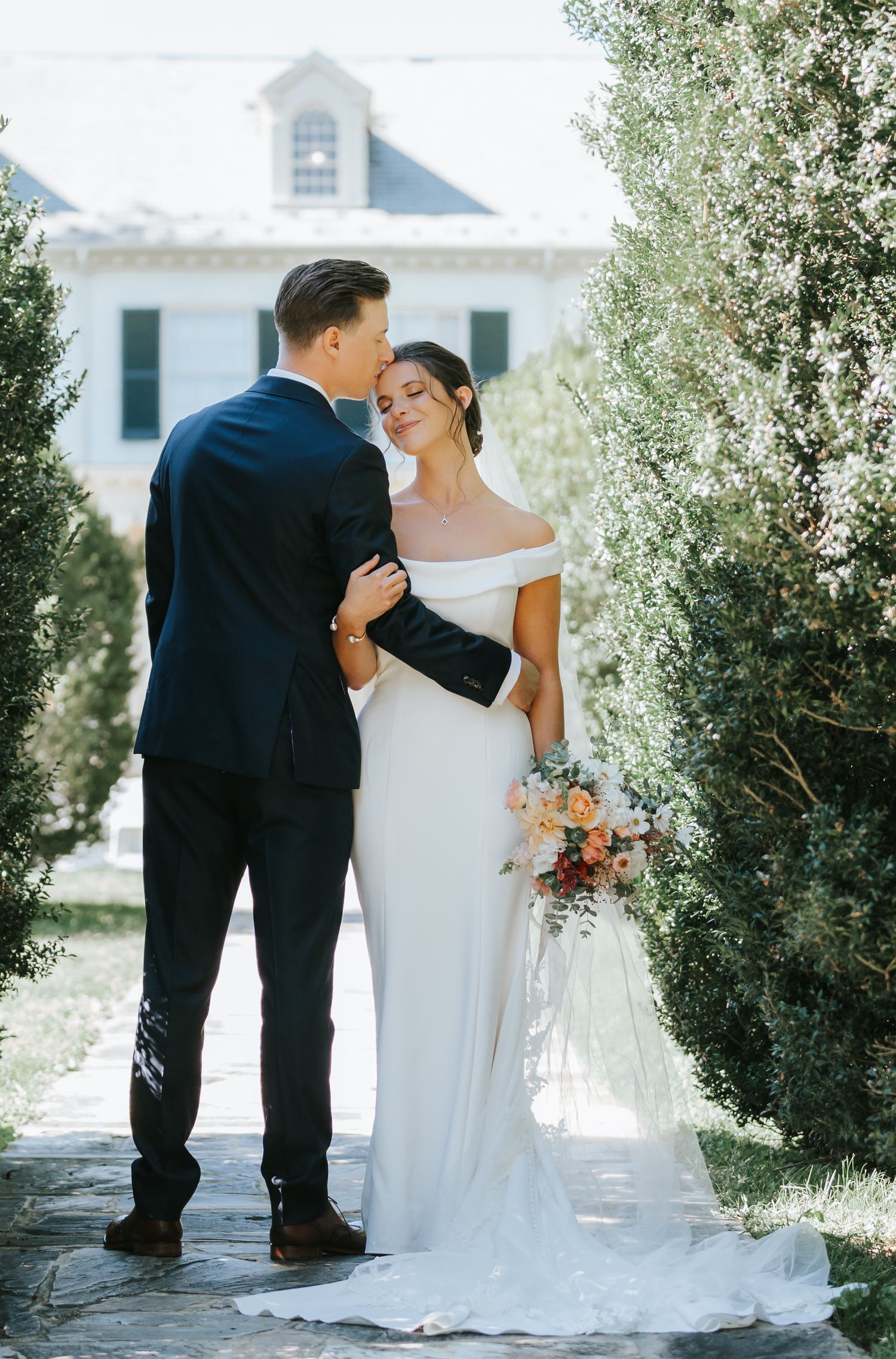 Elegant wedding couple embracing in front of white chapel with flowing bridal gown and floral bouquet.