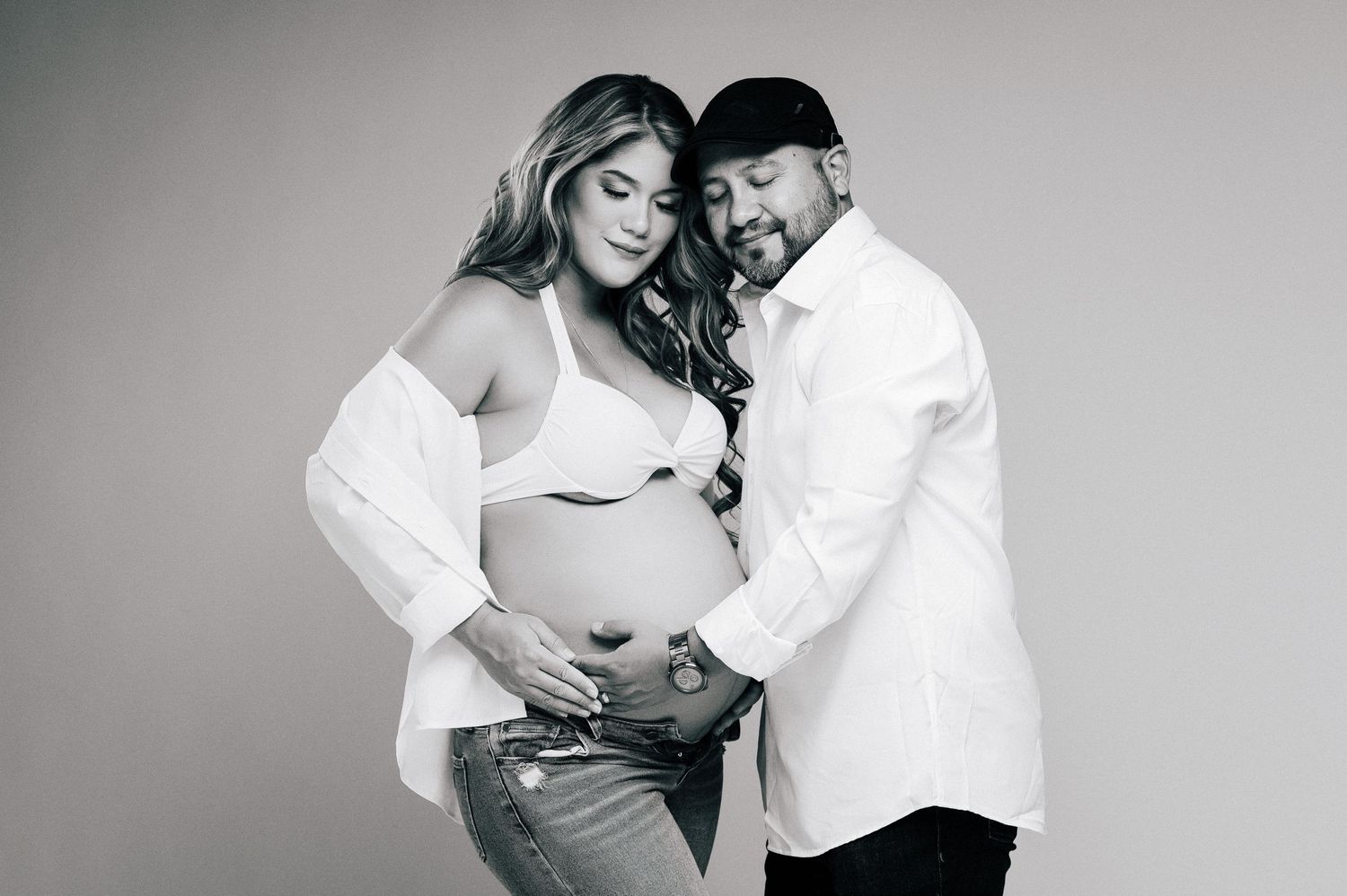 Black and white maternity photo of a couple tenderly cradling a pregnant belly in a studio setting.