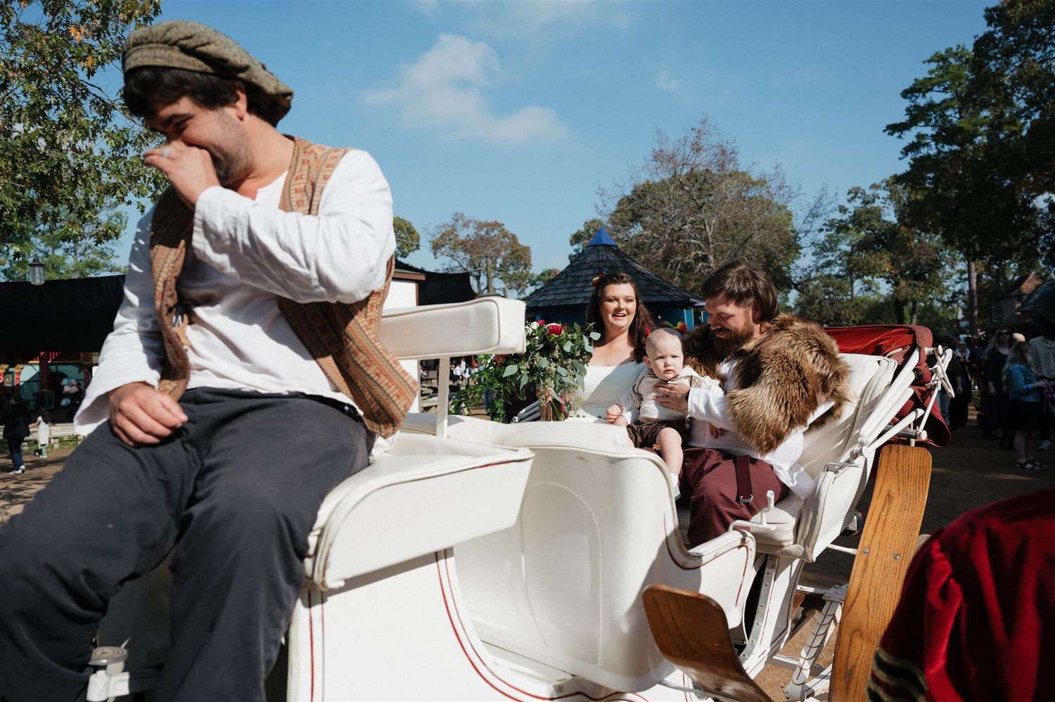 Costumed performers ride in a white horse-drawn carriage at a renaissance faire.