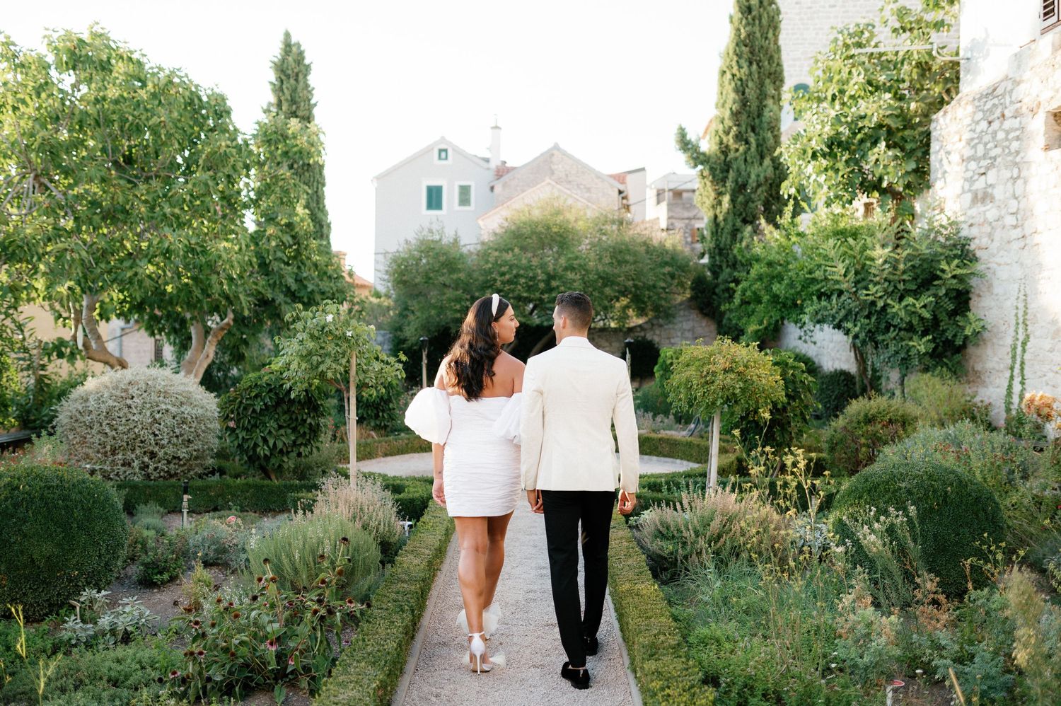Two people walking down a garden path surrounded by lush greenery and cypress trees in a Mediterranean setting.