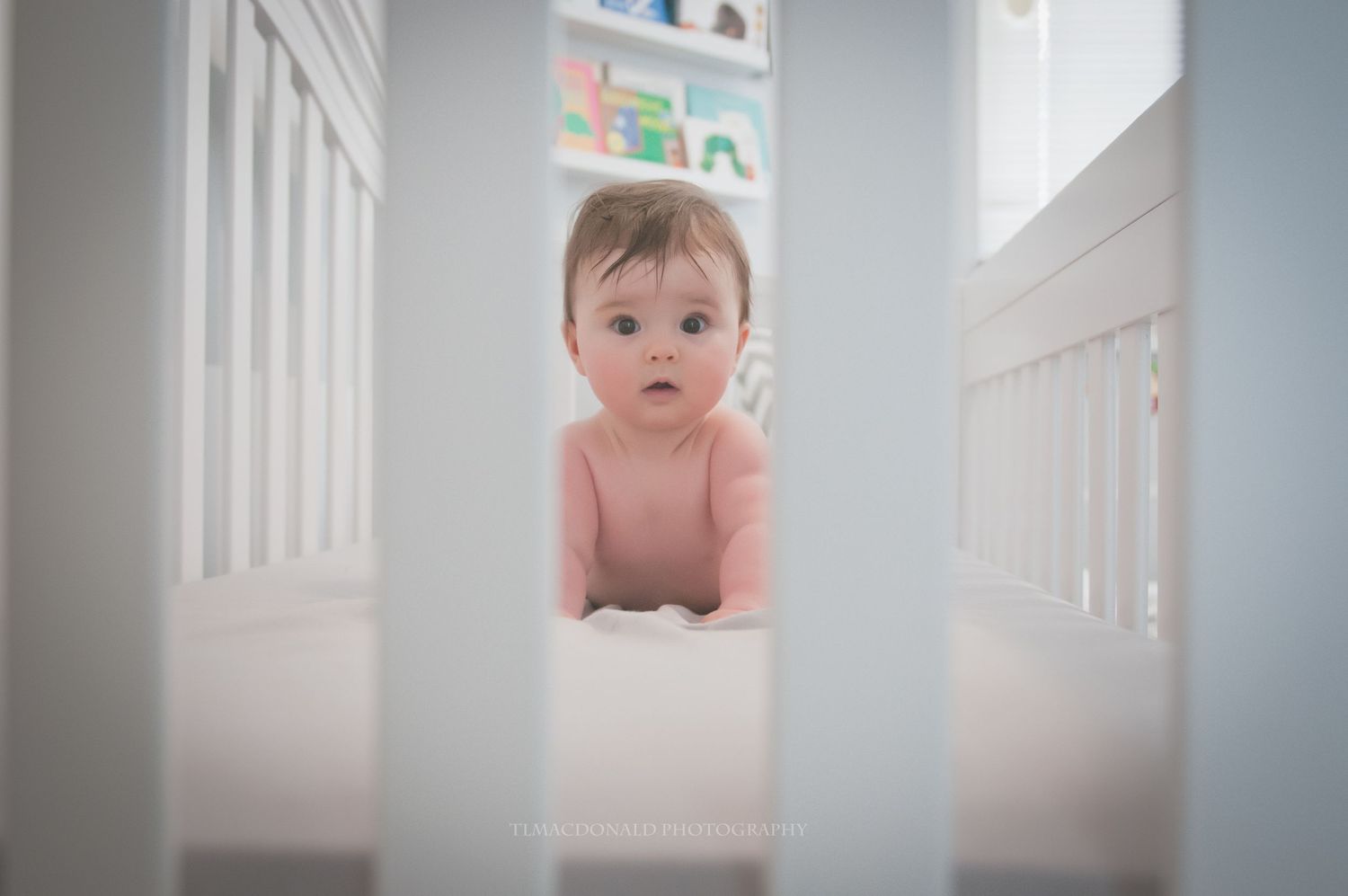 Baby peering through white crib bars with artwork displayed on wall in background.