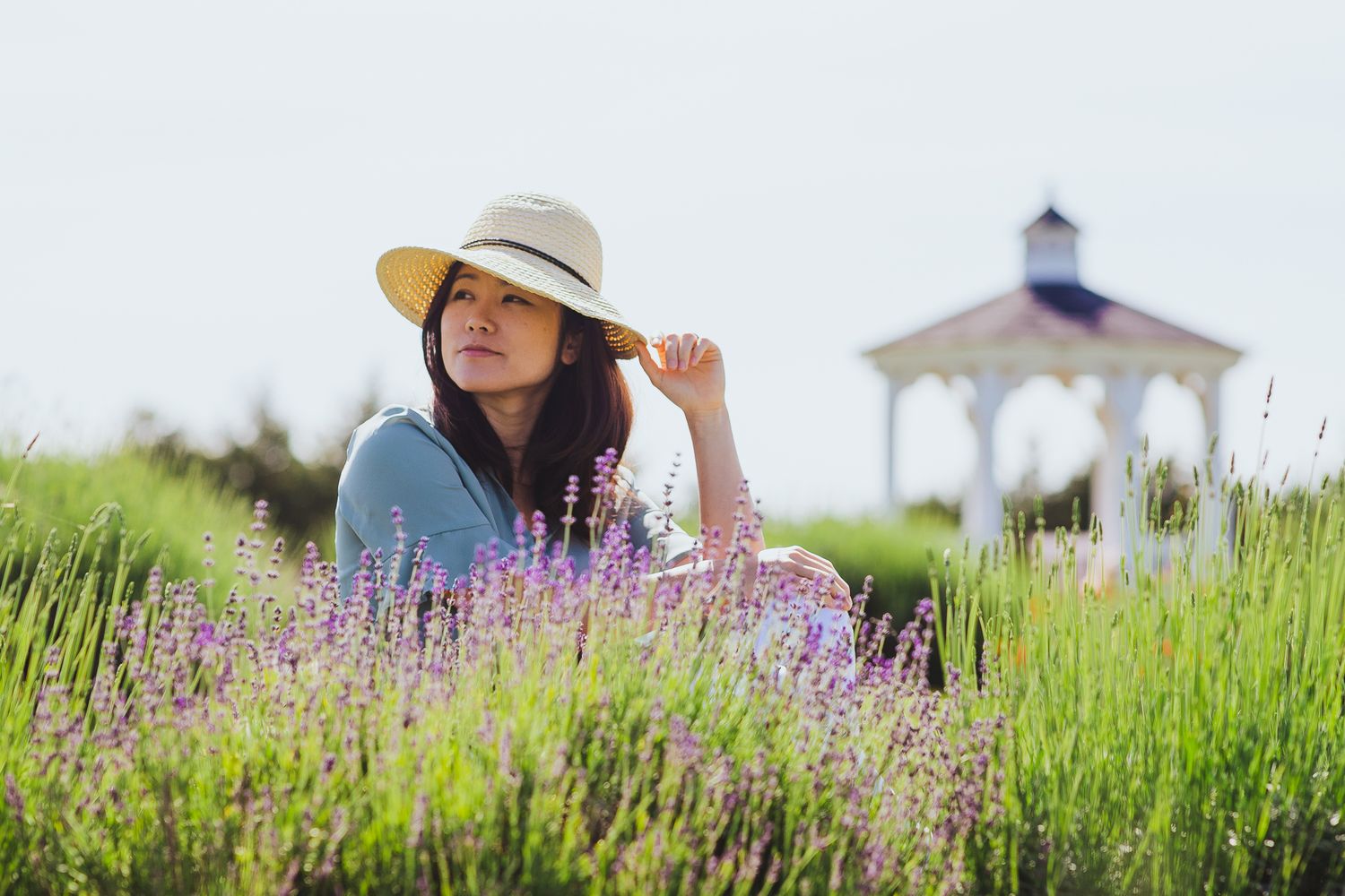 Person in a sunhat poses among purple lavender flowers with a gazebo in the background.