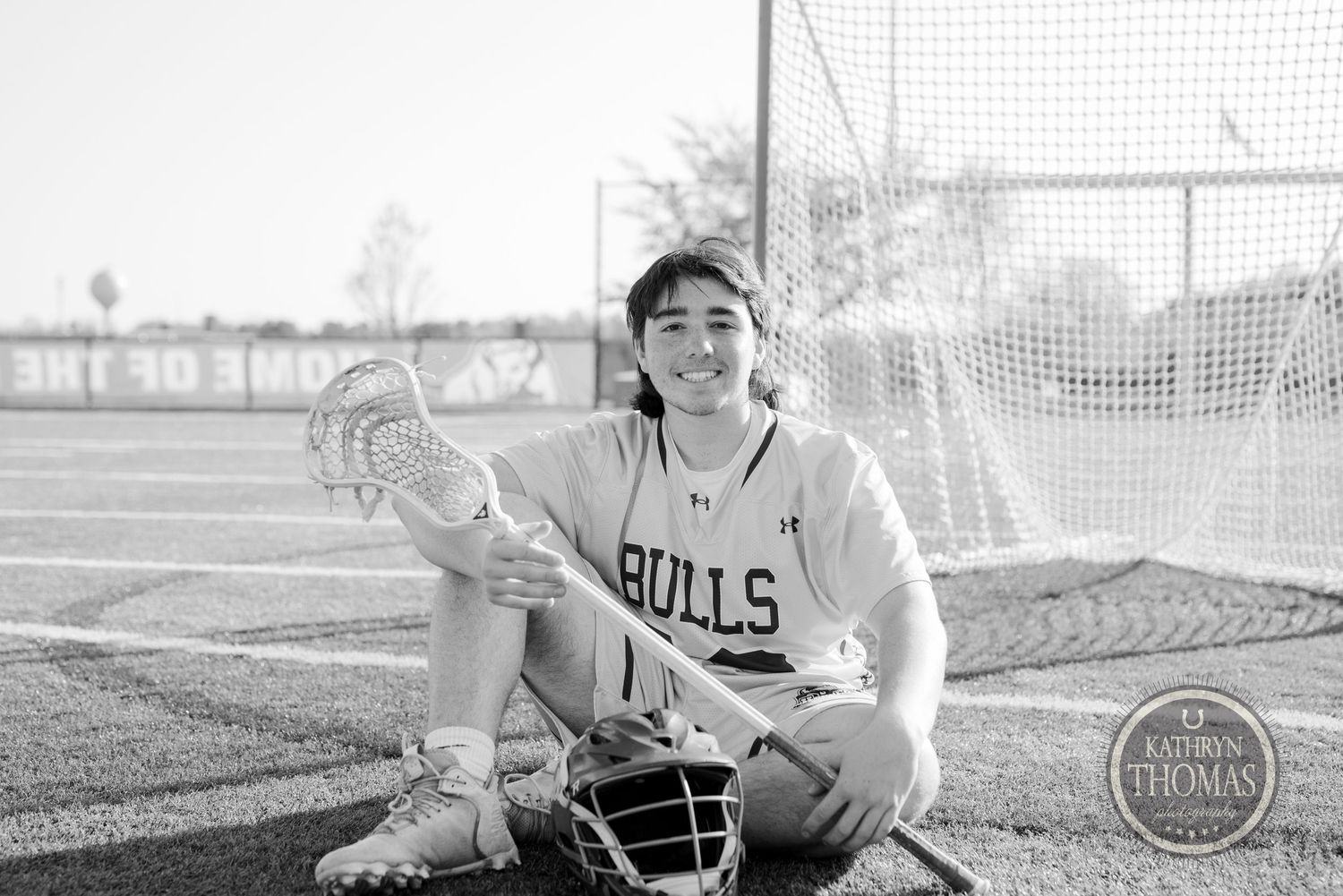 Black and white photo of athlete in Bulls uniform sitting with lacrosse equipment.