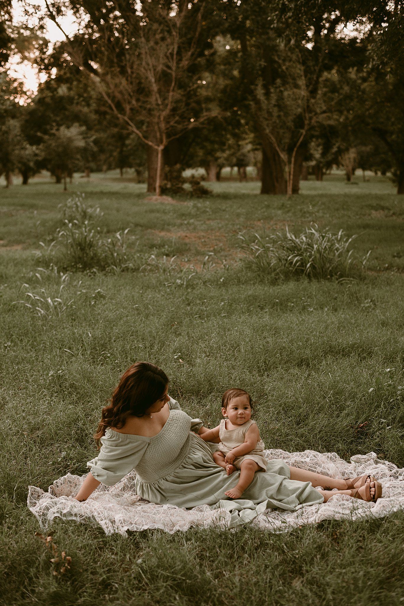A tender outdoor moment on a white blanket under large trees at sunset.