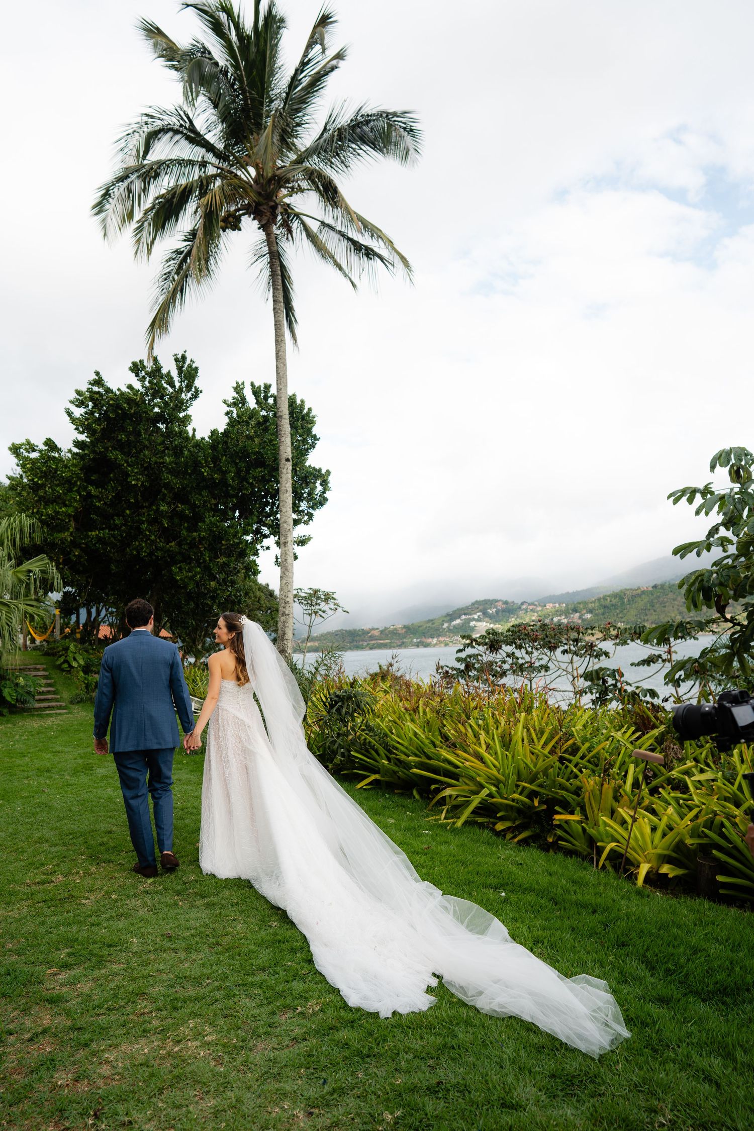 Wedding couple walks through tropical garden with flowing dress train and ocean view.