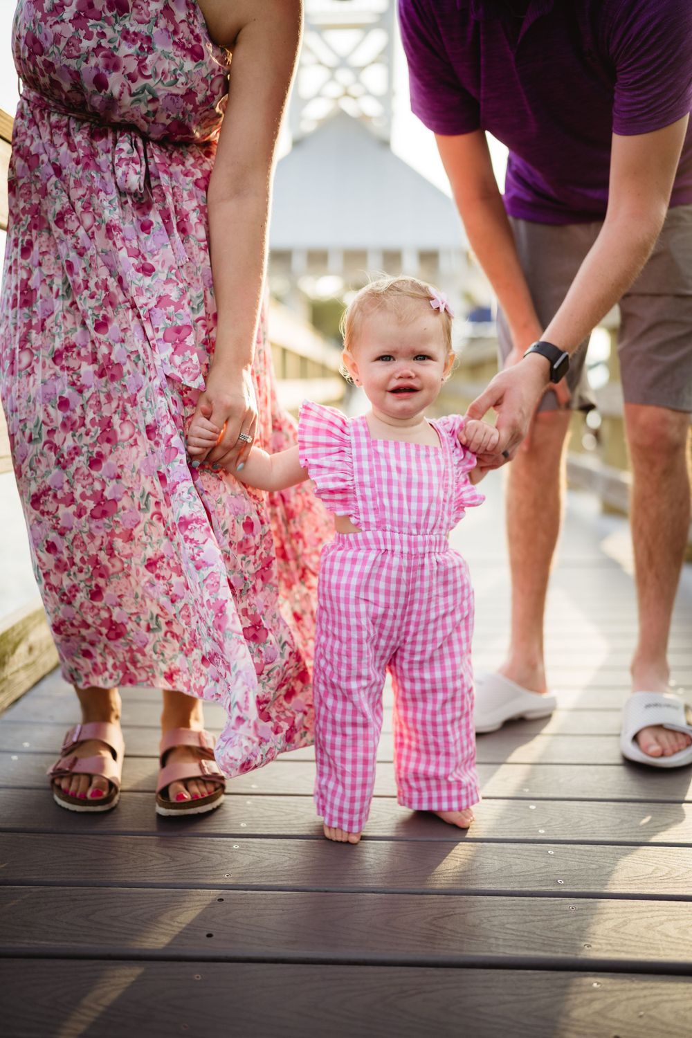 A toddler in pink gingham taking first steps on a wooden boardwalk while holding hands with adults.
