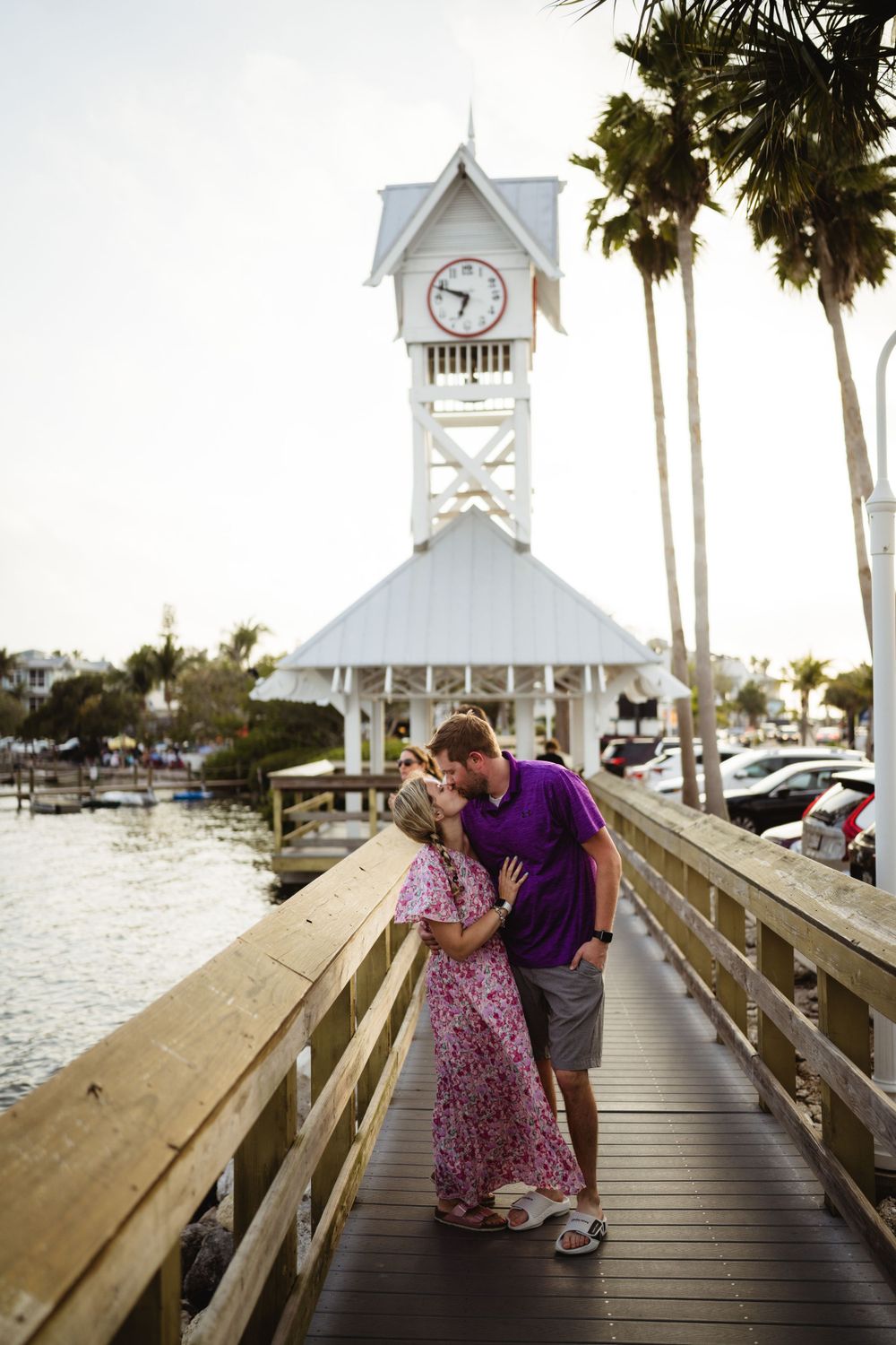 A romantic moment on a pier with a white clock tower and palm trees at sunset.