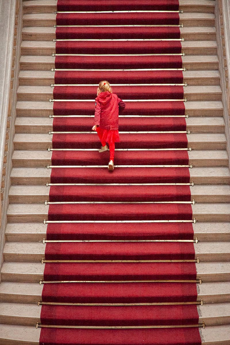 Figure in bright red outfit ascending grand staircase with red carpet.
