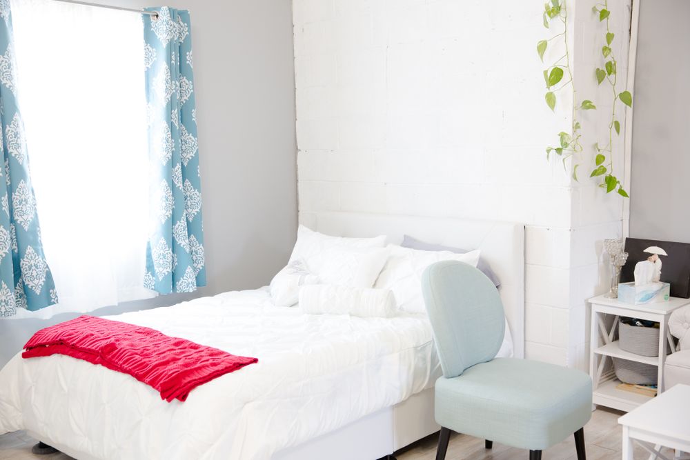 Bright bedroom setup with white bedding, mint accent chair, and blue patterned curtains.