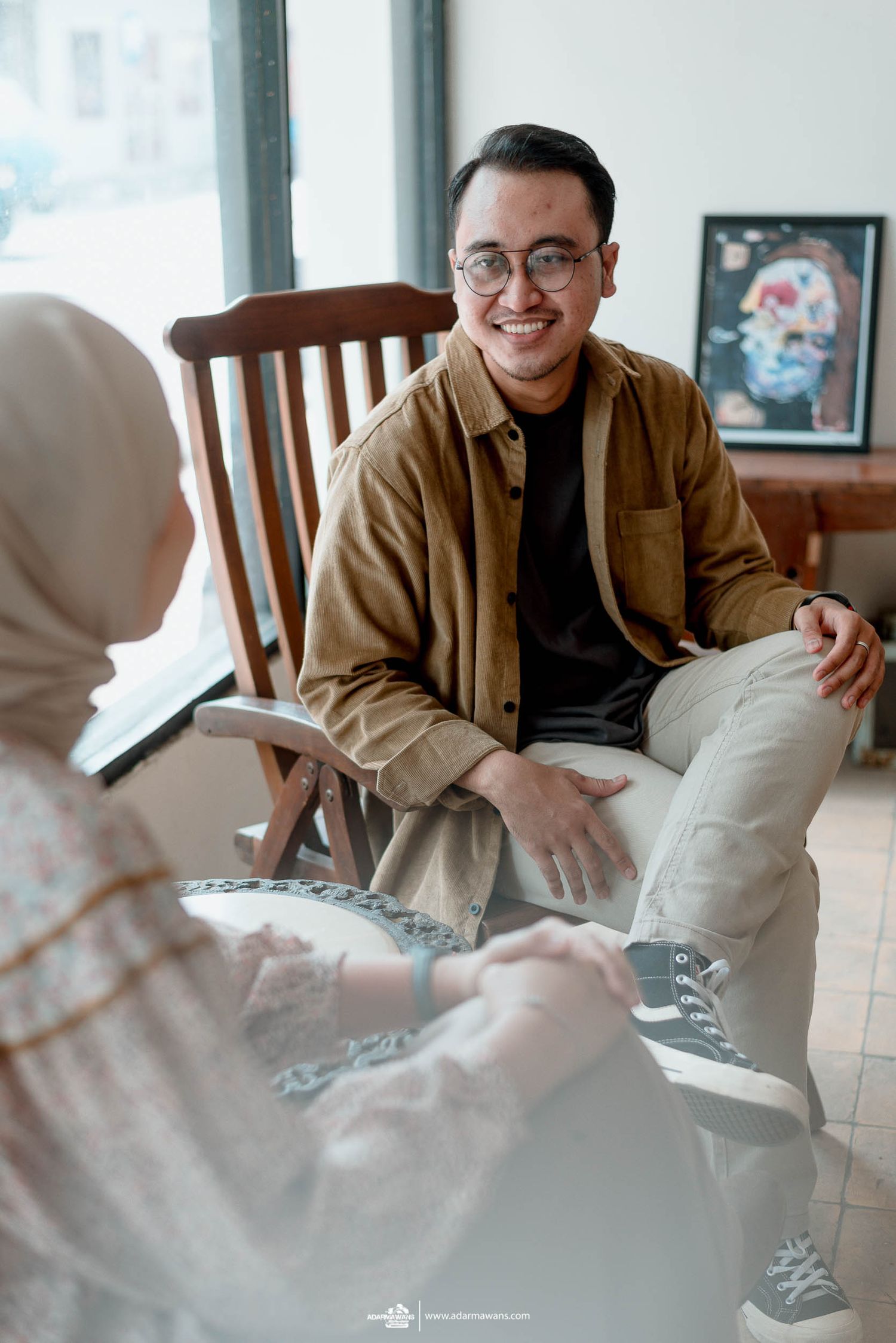 Person in brown jacket and glasses sits casually on wooden chair during conversation.